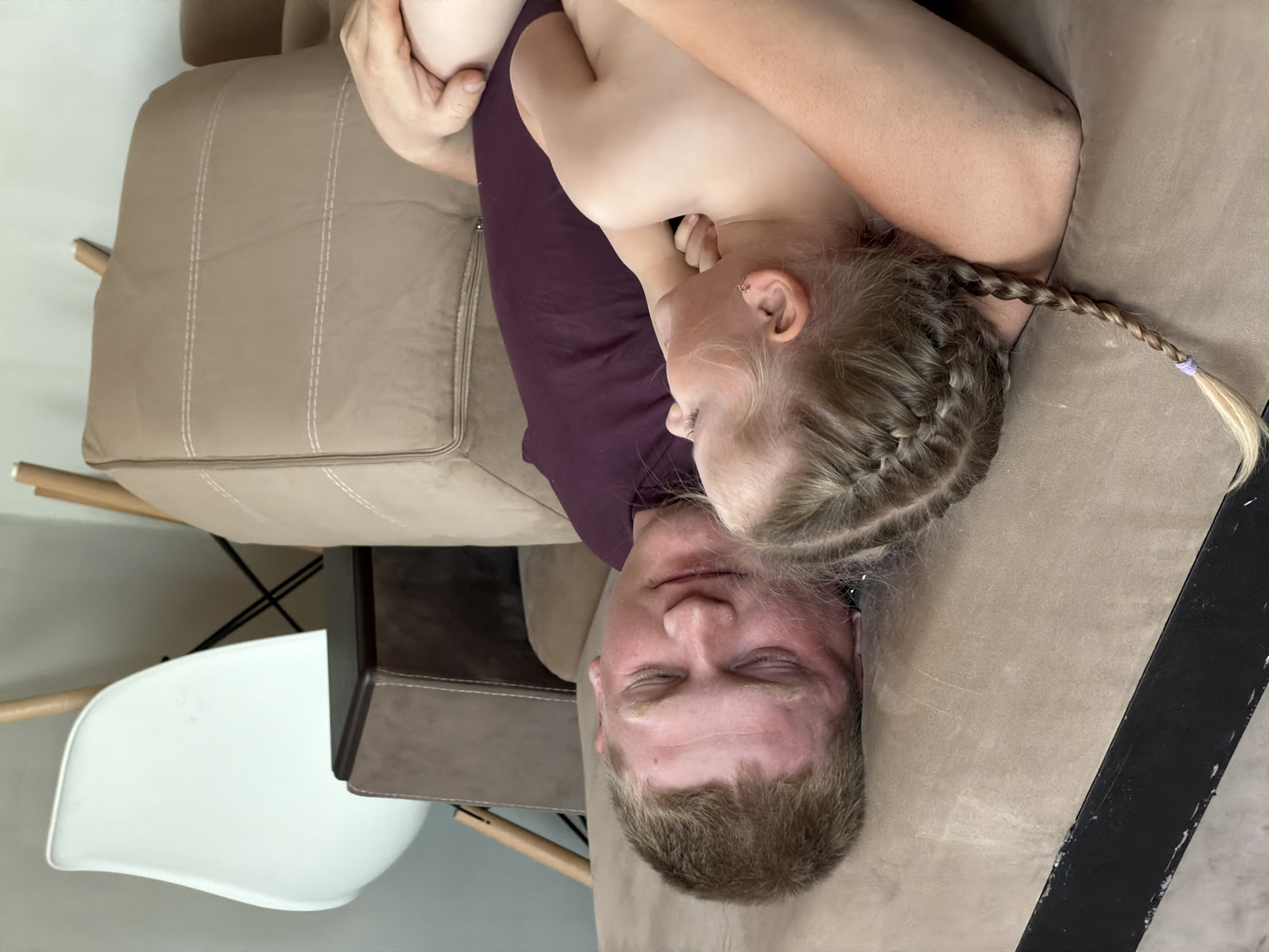 A man and a young girl are shown in the image, which appears to be taken in an indoor setting. The man is lying down with his eyes closed, and the girl is resting on him in an embrace. The girl has blonde hair styled in braids. The man has short, light brown hair. They are both situated on a tan-colored sofa. A white chair with wooden legs is visible in the background. The image does not offer enough information to determine whether the location is in Chaikovsky, Russia.