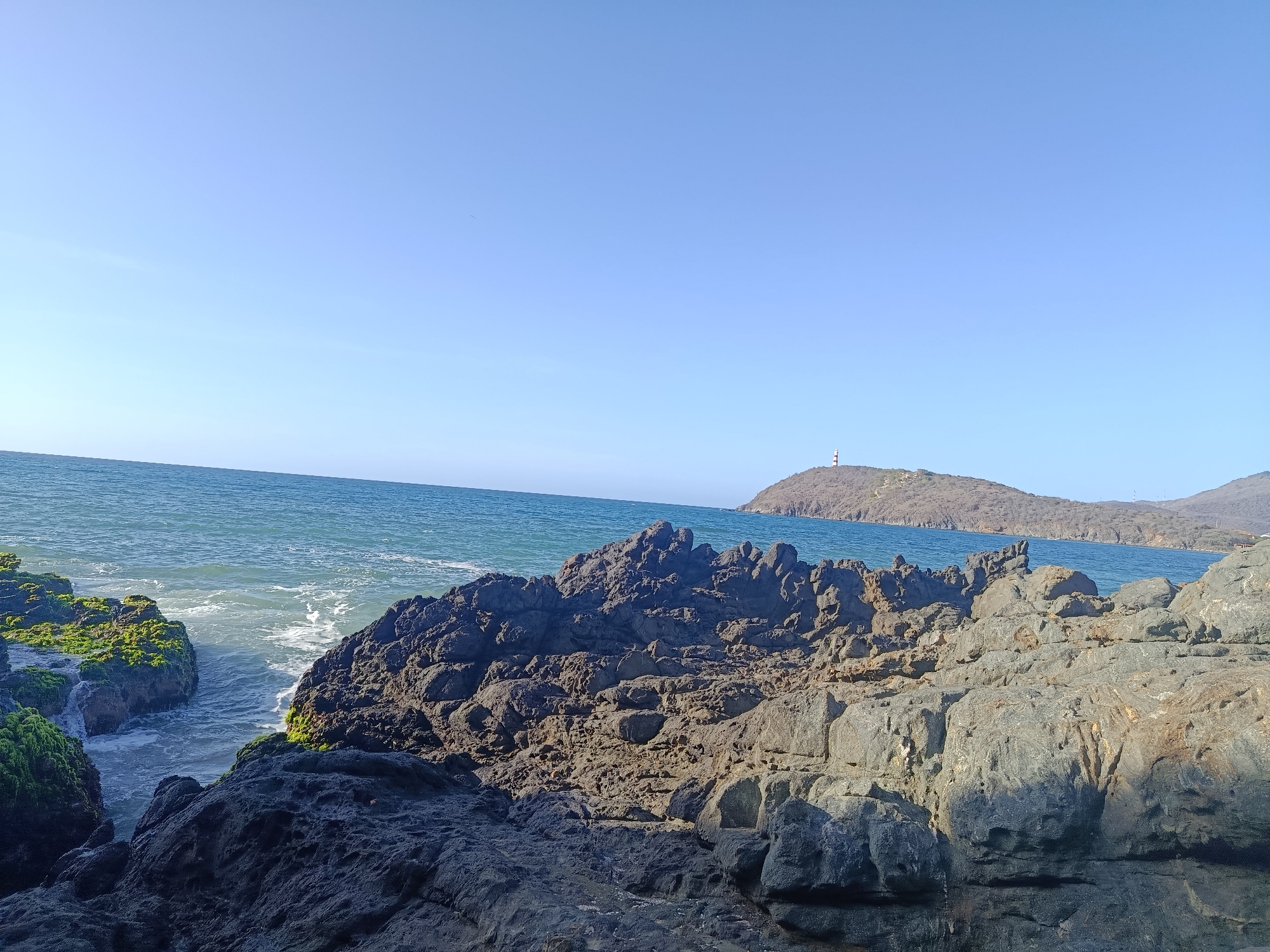 A rocky coastline with waves crashing against the shore is seen under a clear blue sky. In the distance, a hill with a lighthouse on top overlooks the ocean. The foreground is dominated by dark, rugged rocks, some of which are covered in green moss. The ocean is a deep blue, with whitecaps visible as the waves roll in. The overall scene depicts a natural coastal landscape. The location is Pedro González, Venezuela.