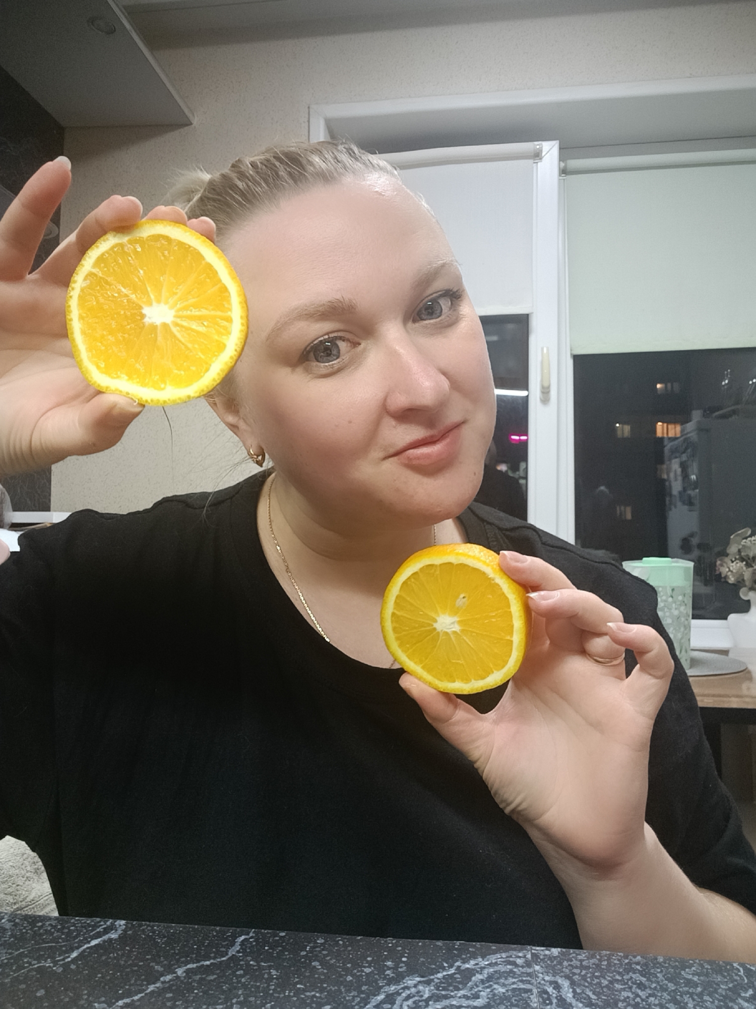 A woman is holding two orange halves in front of her. She is fair-skinned with blonde hair pulled back in a ponytail. She is wearing a black shirt and a gold chain. The background appears to be a kitchen or dining area with a window overlooking a cityscape at night. Based on metadata, the image was taken in Chaikovsky, Russia.