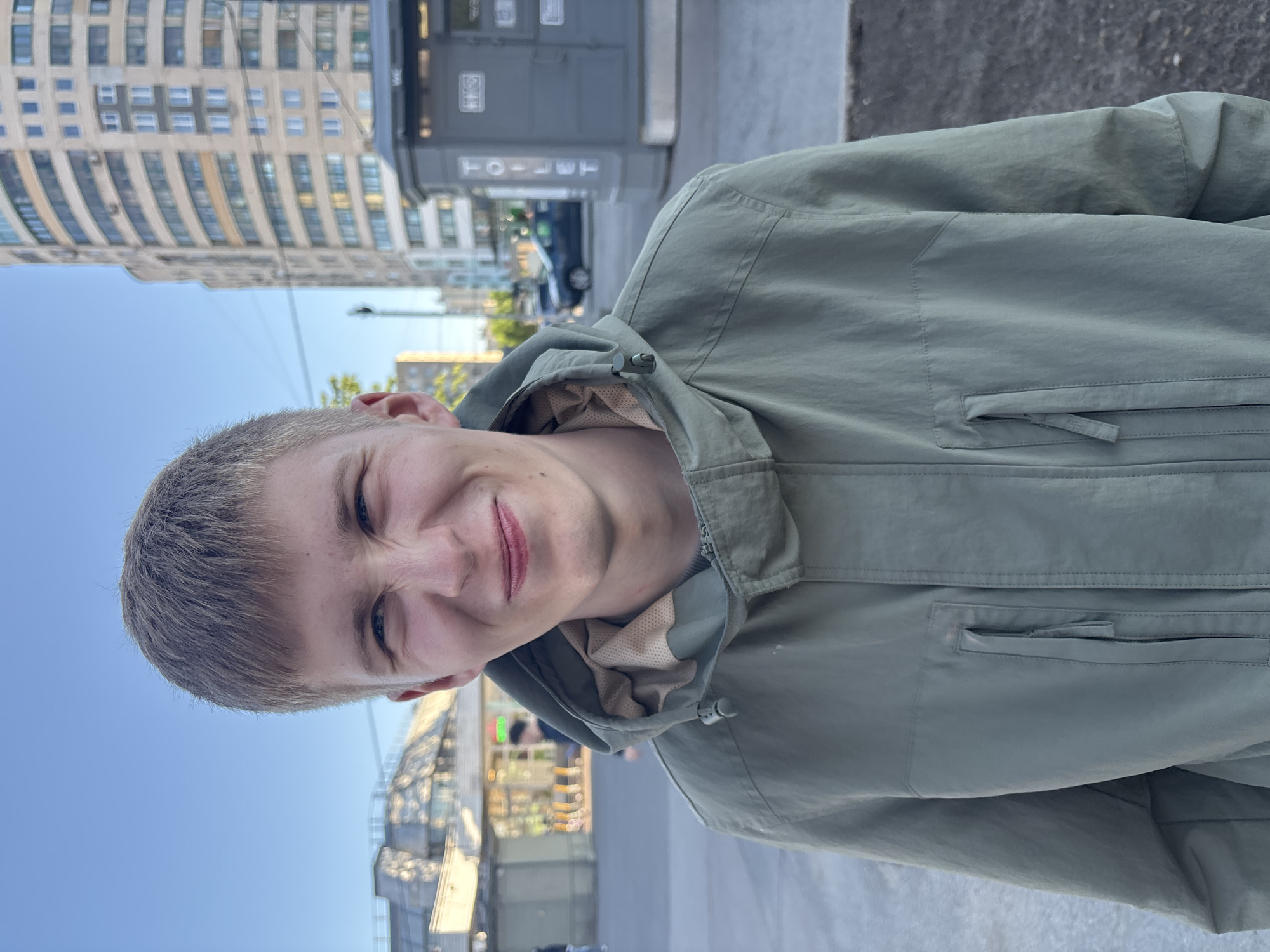 A young man with short, light brown hair is posing for a photo outdoors, wearing a light green hooded jacket. The background includes a tall building with many windows, and a gray utility structure labeled "TOILET". In the distance, blurred vehicles and other buildings can be seen. The sky is blue. The location appears to be an urban environment, likely within Vsevolozhsk, Russia.