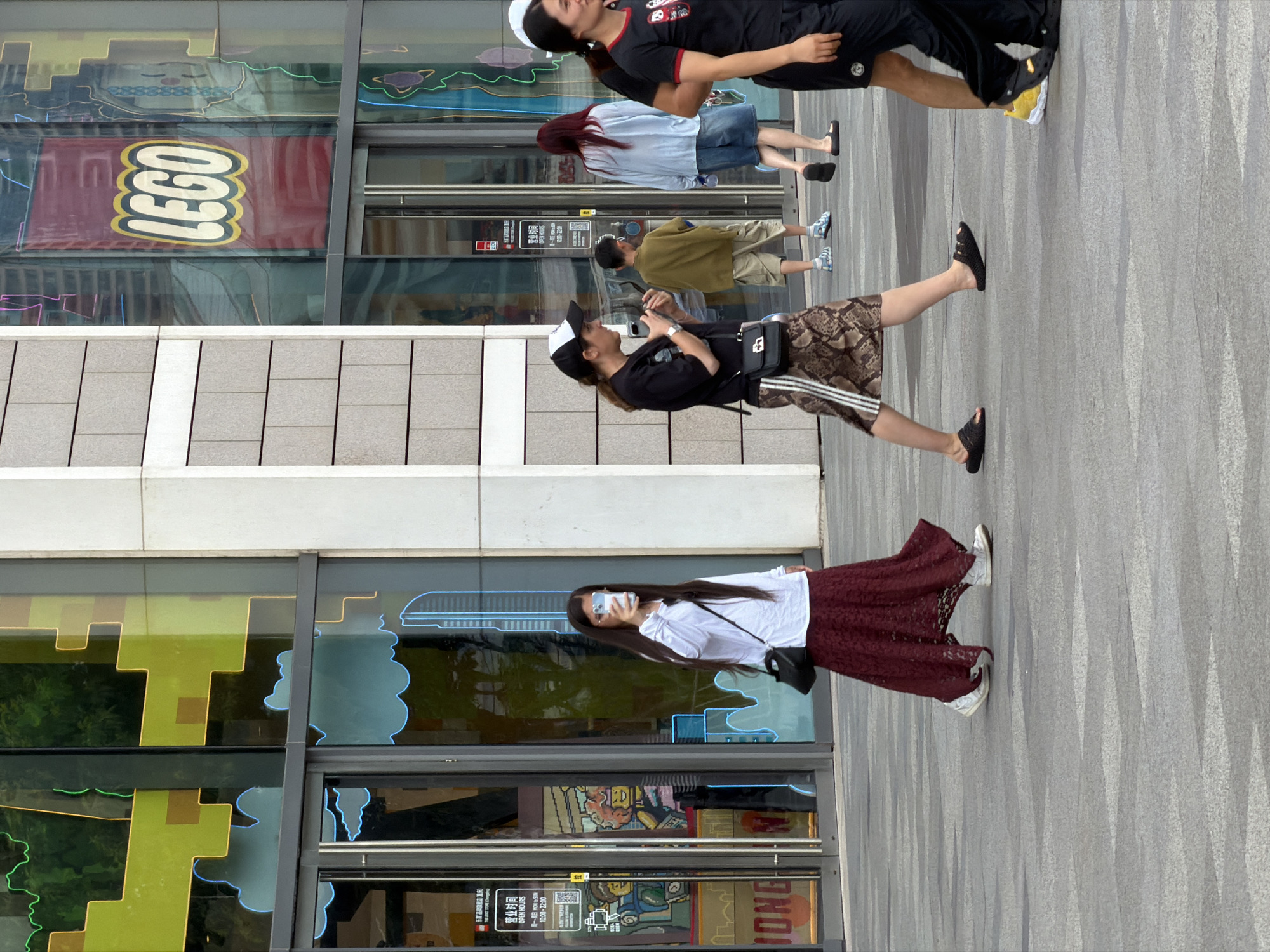 In Yuzhong District, China, a street scene unfolds in front of a LEGO store. The store's facade features large glass windows displaying colorful graphics, including what appears to be a pixelated design and cloud shapes. A prominent red banner with the white LEGO logo is visible on the upper right side of the building. Several people are walking on the paved ground in front of the store.
On the left, a woman with long dark hair is walking away from the camera, holding a smartphone in her right hand. She is wearing a white long-sleeved shirt, a dark bow at her neck, and a long, flowing dark red lace skirt. She has a black crossbody bag with a silver clasp and is wearing white sneakers.
To her right, a woman wearing a black t-shirt, a white and black cap, and a patterned skirt with stripes on the side is walking towards the camera, looking at her smartphone. She is carrying a black crossbody bag with a silver clasp and wearing dark slides.
Behind them, a young boy in a green shirt and beige shorts walks away from the camera. Further to the right, a woman with long red hair in a light blue shirt and denim shorts walks away. To her right, a woman in a black t-shirt with red trim and a panda graphic on the chest walks towards the camera, smiling. Another person in black pants and a black shirt is also visible. The ground is paved with gray tiles arranged in a patterned walkway.