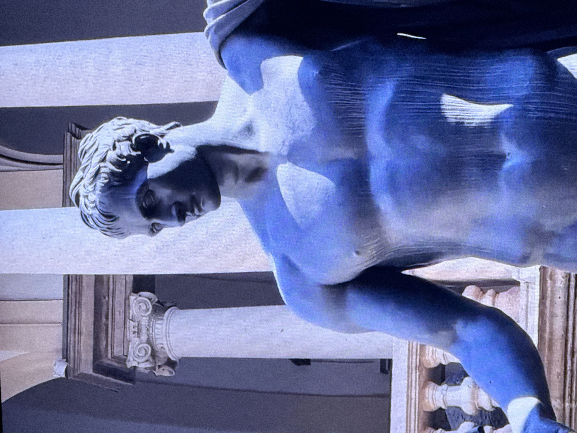 The image shows a close-up of a blue-toned statue of a muscular male figure. The statue is depicted from the chest up, with its head turned to the left. The background features classical architecture, including fluted columns with Corinthian capitals and a balustrade. The lighting creates strong shadows, highlighting the musculature of the statue. There is no indication of the statue's location in Dubai, United Arab Emirates.