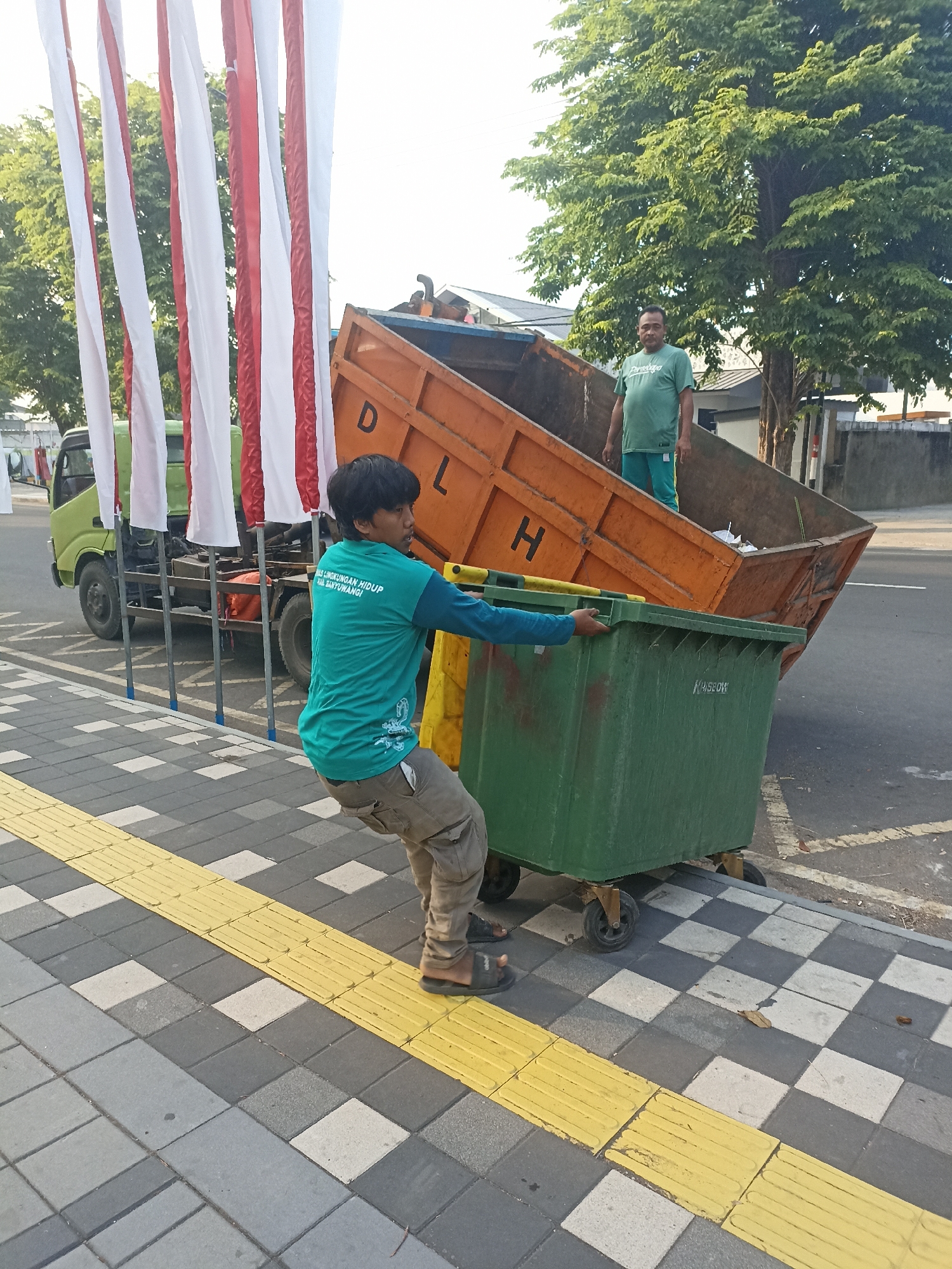 Two men are engaged in waste disposal activities on a street in Kelurahan Kepatihan, Indonesia. In the foreground, a man wearing a teal t-shirt and khaki pants is pushing a large green trash bin on wheels. He is positioned on a sidewalk paved with grey and white square tiles, with a tactile yellow paving strip running alongside it. In the background, an orange dumpster truck is parked, its bed tilted upwards. A second man, wearing a green t-shirt and green pants, stands inside the truck's bed. Red and white flags are visible on poles to the left of the truck. The scene appears to be during daylight hours, with trees and buildings in the background.