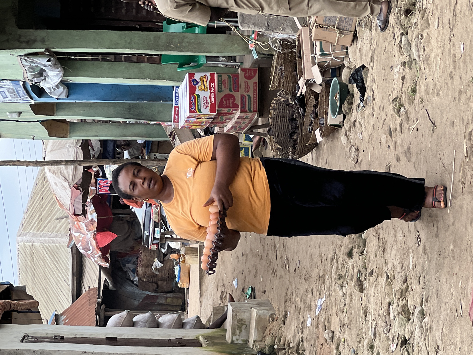 A woman in a bright orange t-shirt and black pants walks through a market in Oko Amakom, Nigeria, holding a carton of eggs. She is wearing sandals. The market appears to be outdoors, with stalls and buildings made of corrugated metal and concrete. Various goods are visible, including sacks of produce, baskets of dried fish, and boxes of instant noodles. The ground is uneven and covered with dirt, rocks, and some litter. In the background, other market-goers and a car are visible. The sky is overcast.