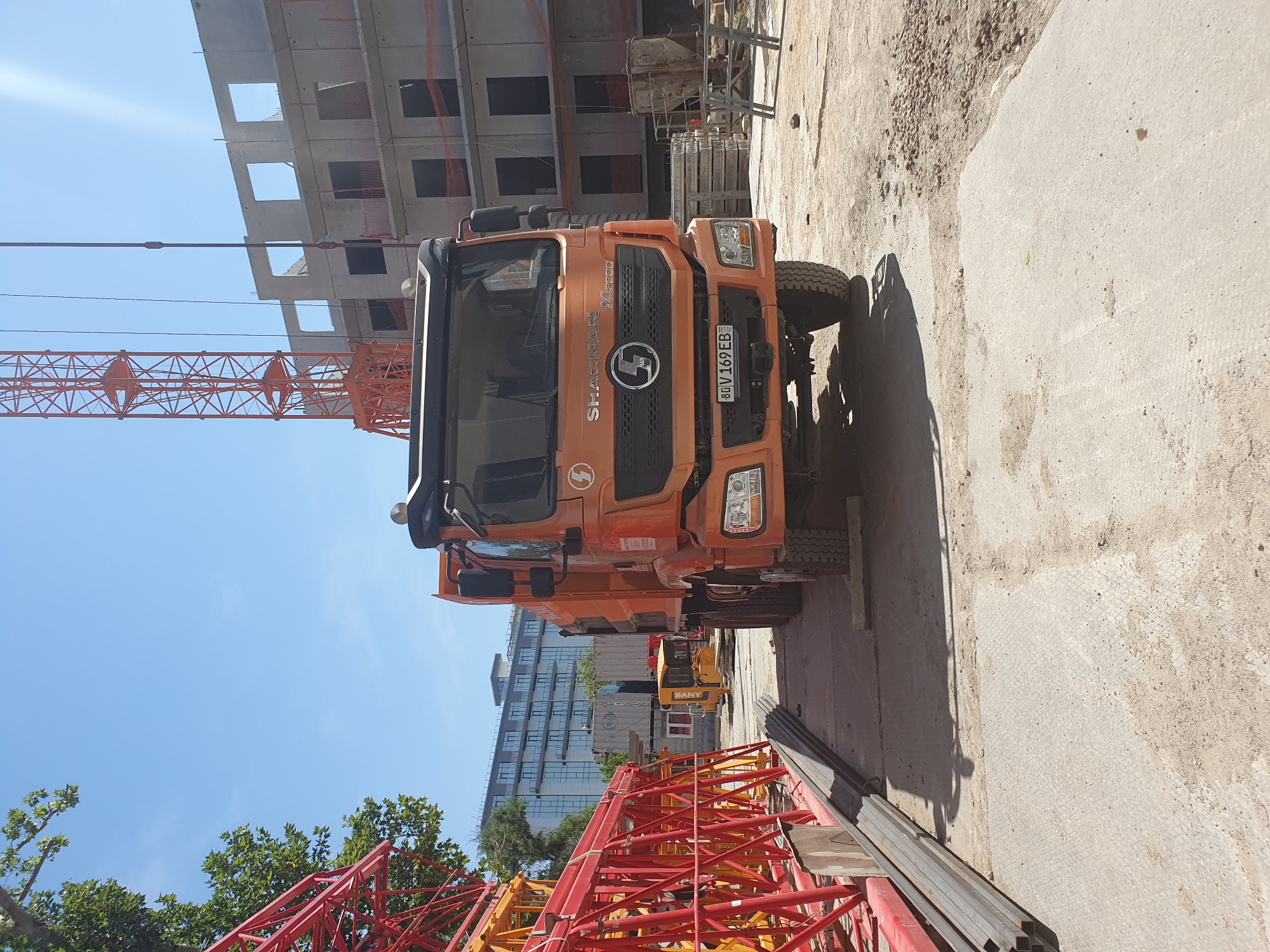 An orange Shacman M3000 dump truck is parked on a dirt and concrete surface in what appears to be a construction site in Tashkent, Uzbekistan. The truck is facing forward, with its front grille prominently displayed, featuring the Shacman logo and license plate "80 V 169 EB". In the background, a multi-story building under construction with visible concrete framing and empty window openings stands under a clear blue sky. A tall orange tower crane is positioned to the left of the building. In the foreground, to the left of the truck, are stacked metal beams and a section of a red construction crane. A yellow mini-excavator is visible further into the site behind the truck. The environment suggests an active construction zone.