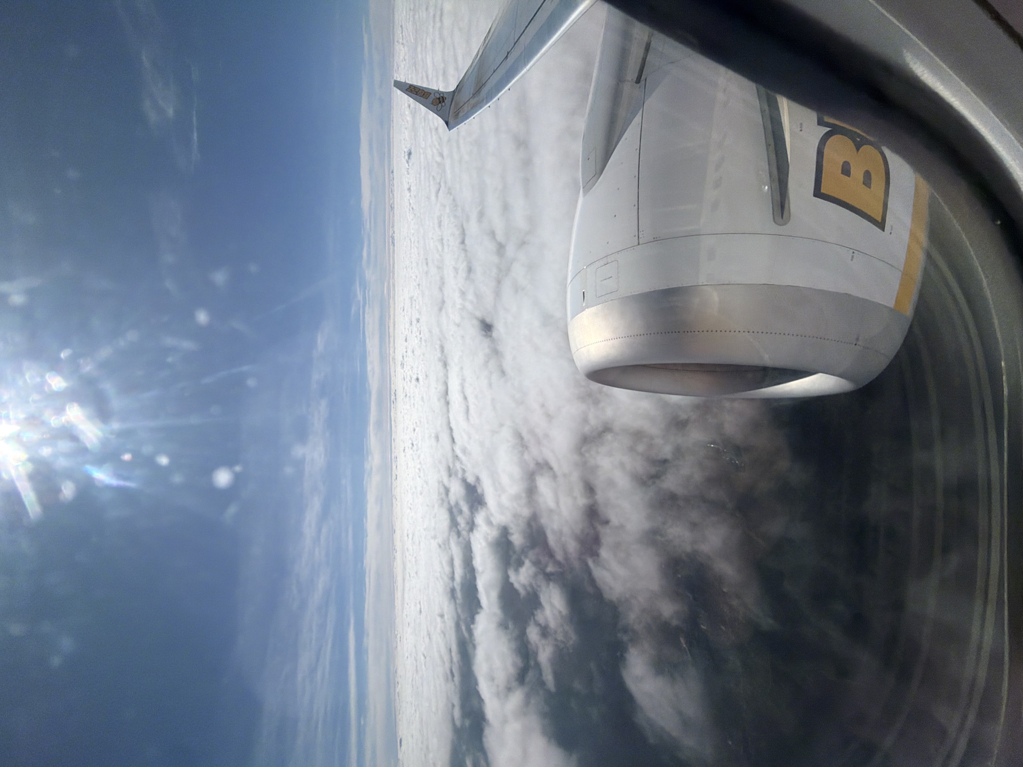 The view from an airplane window shows a bright blue sky on the left and a sea of white clouds below. Part of the airplane's wing and engine is visible on the right. The engine has a stylized "B" logo on it. The image was taken from an aircraft, not in the city of Przeginia Duchowna, Poland.