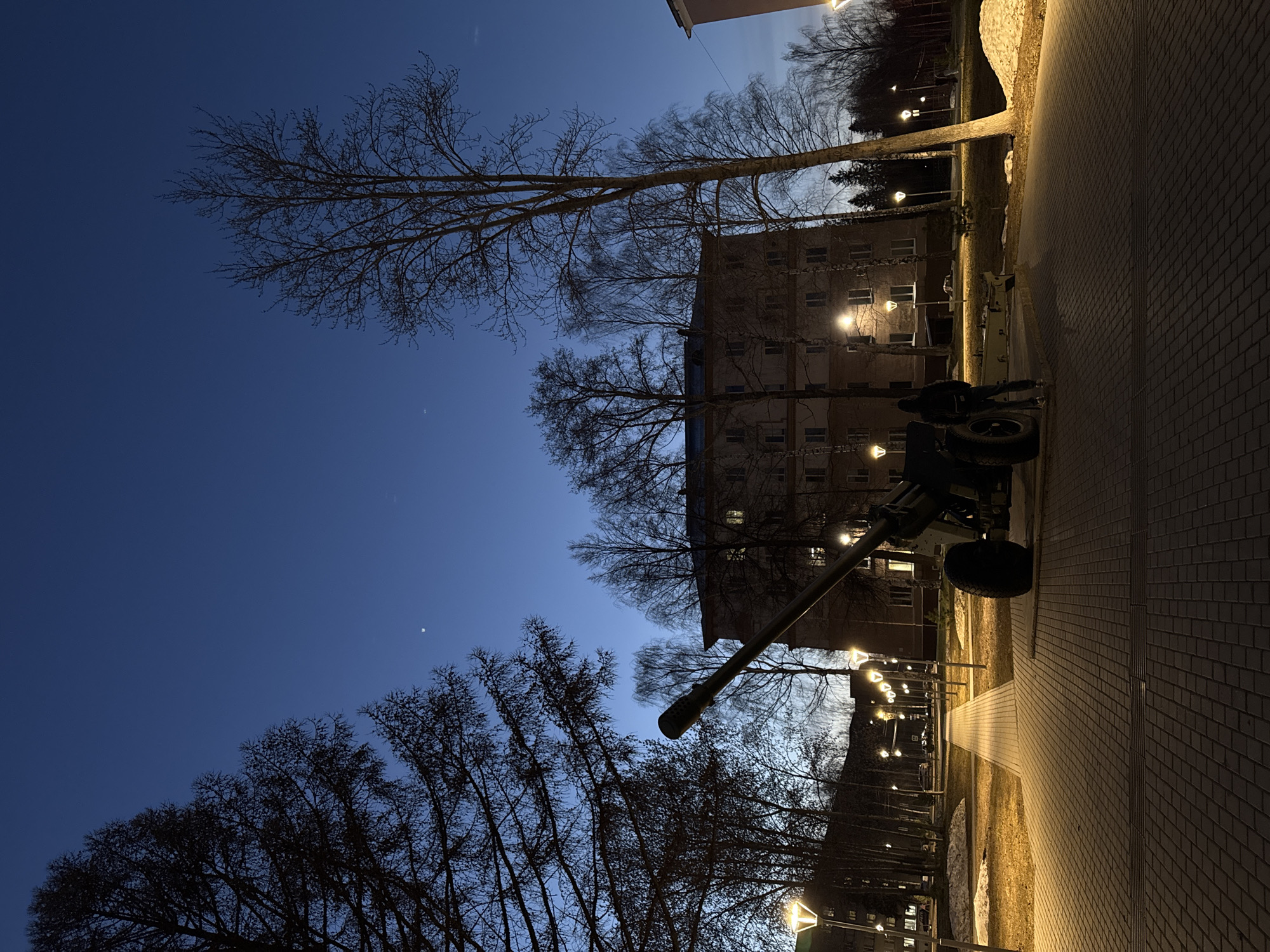 A ground-level, nighttime shot of an anti-tank gun memorial in Izhevsk, Russia. The monument, positioned to the right of frame, stands near a brick walkway illuminated by streetlights, against a backdrop of bare trees and a multi-story residential building with lit windows. The sky is a dark blue, with a crescent moon visible at the bottom-left.
