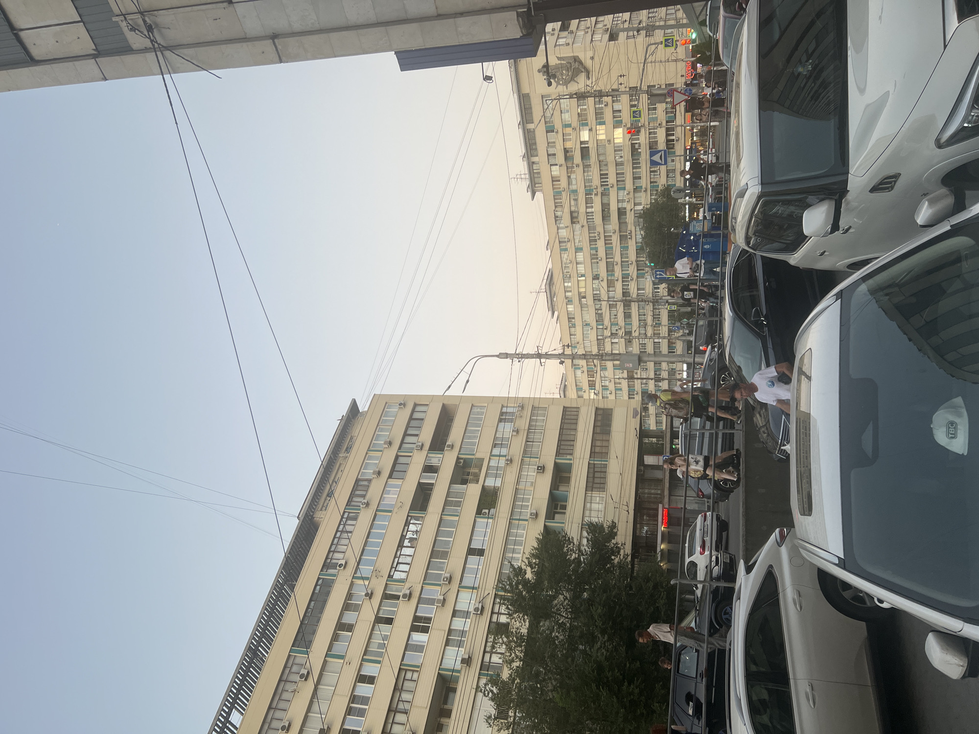 This is a photograph taken from a street-level perspective in Volgograd, Russia, likely during daylight with a clear sky, suggesting it's either early morning or late afternoon due to the soft light. The scene is urban, characterized by multi-story apartment buildings and heavy traffic.
The foreground is dominated by the hoods and windshields of several cars. A white SUV, possibly a Lexus, is prominent on the right, its side visible. To its left, another white vehicle, likely also an SUV, is partially visible. In the center, a dark-colored sedan is parked.
In the middle ground, a street runs horizontally across the image, lined with more cars. A group of people are walking on a sidewalk next to the street, some appearing to be pedestrians or shoppers. One person in a white t-shirt is visible near the white SUV in the foreground. Further down the street, traffic lights and street signs are visible, indicating an intersection. Several tall, light-colored apartment buildings with numerous windows and air conditioning units are the dominant structures in the background. Some of these buildings appear to be connected by overhead wires, likely for power or telecommunications.
On the left side of the image, a large, dark green tree provides a natural element against the urban backdrop. A person in a light-colored shirt and pants stands near the tree, seemingly observing the street. The sky above is a pale blue, with thin, dark lines of wires crisscrossing it. The overall impression is of a busy, everyday scene in a Russian city.
