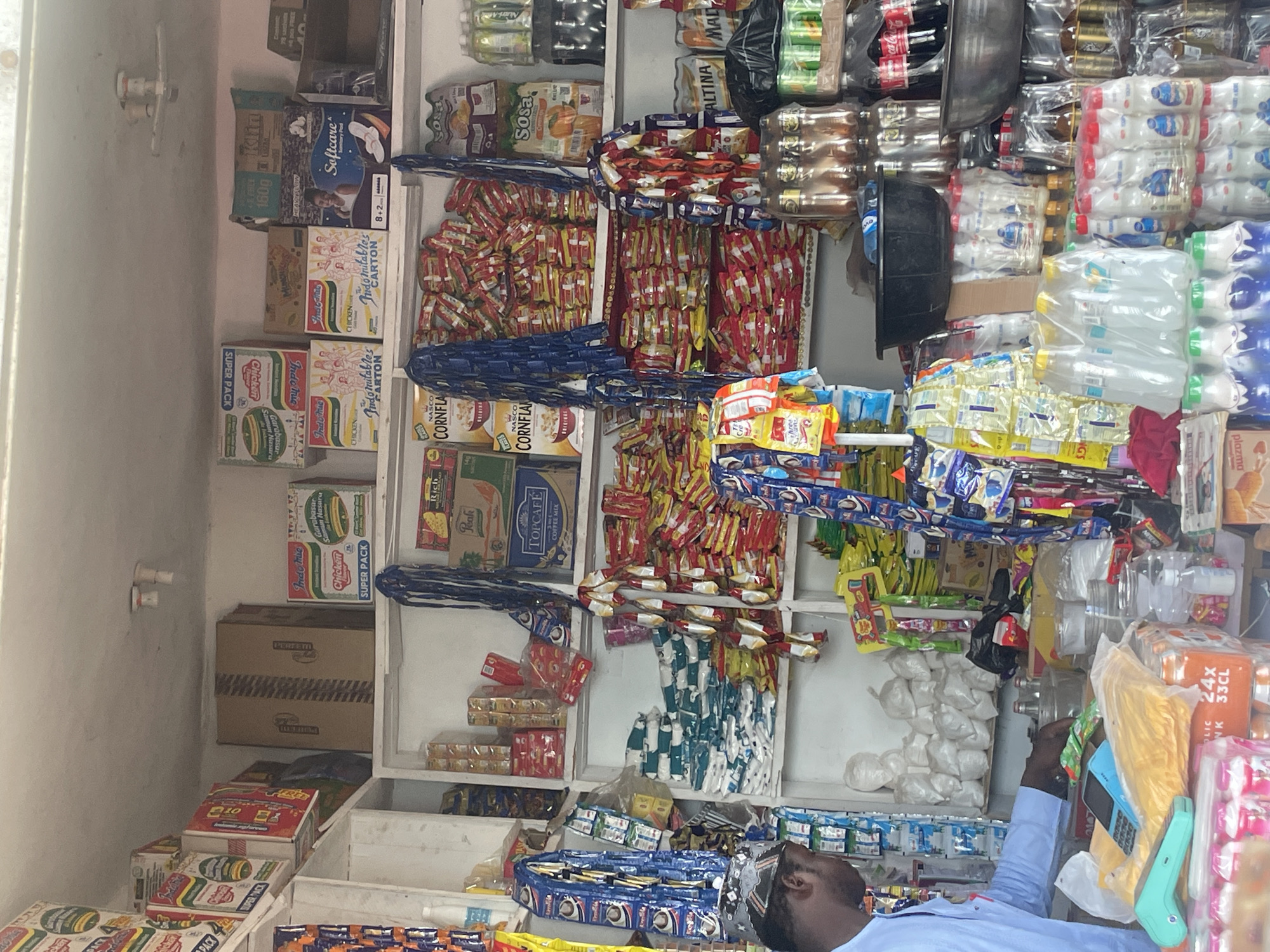 This is a medium shot photograph of the interior of a small convenience store, likely a kiosk, located in the city of Bauchi, Nigeria. The store is densely packed with various products displayed on shelves and hanging from the ceiling. The overall impression is one of abundance and variety, typical of many small retail businesses in Nigeria. **People:** A single male individual is visible in the left portion of the frame. He is wearing a light blue long-sleeved shirt and is facing away from the camera, towards the shelves. His right hand is extended, seemingly reaching for or interacting with an item on a lower shelf. He appears to be a shopkeeper or an employee. **Objects and Products:** The shelves are filled with a wide array of packaged goods. Prominent items include: * **Food and Beverages:** Numerous packets of instant noodles (Indomie is clearly visible), cornflakes, biscuits, coffee mix, and bottled drinks (soda and what appear to be beverages in plastic bottles). There are also bags of what seem to be white powdery substances on the upper shelves. * **Household Items:** Packs of sanitary pads (Softcare is visible) are also displayed. * **Electronics/Payment Device:** A blue and grey mobile phone and a turquoise card reader are placed on a yellow plastic bag on the counter in the foreground. * **Containers:** A large black plastic basin is placed on a shelf, and a metal bowl is visible to its right. Several glass bottles and jars containing liquids are also present. * **Packaging:** A significant portion of the products are in brightly colored packaging, with a lot of red, yellow, and blue. Many items are sold in multi-packs or individual sachets. **Environment:** The store appears to be a small, enclosed space with a low ceiling. The walls and shelves are predominantly white, showing some signs of wear and tear. The lighting seems to be a mix of overhead fluorescent lights and natural light filtering in from the front of the store, which is not visible in the image. The overall arrangement suggests a functional, perhaps somewhat cluttered, retail environment. The density of products indicates a busy trade. **Actions and Interactions:** The main action is the shopkeeper interacting with the items on the shelf. The presence of the mobile phone and card reader suggests that payment transactions are a part of the store's operation. **Location Context:** The mention of Bauchi, Nigeria, provides context