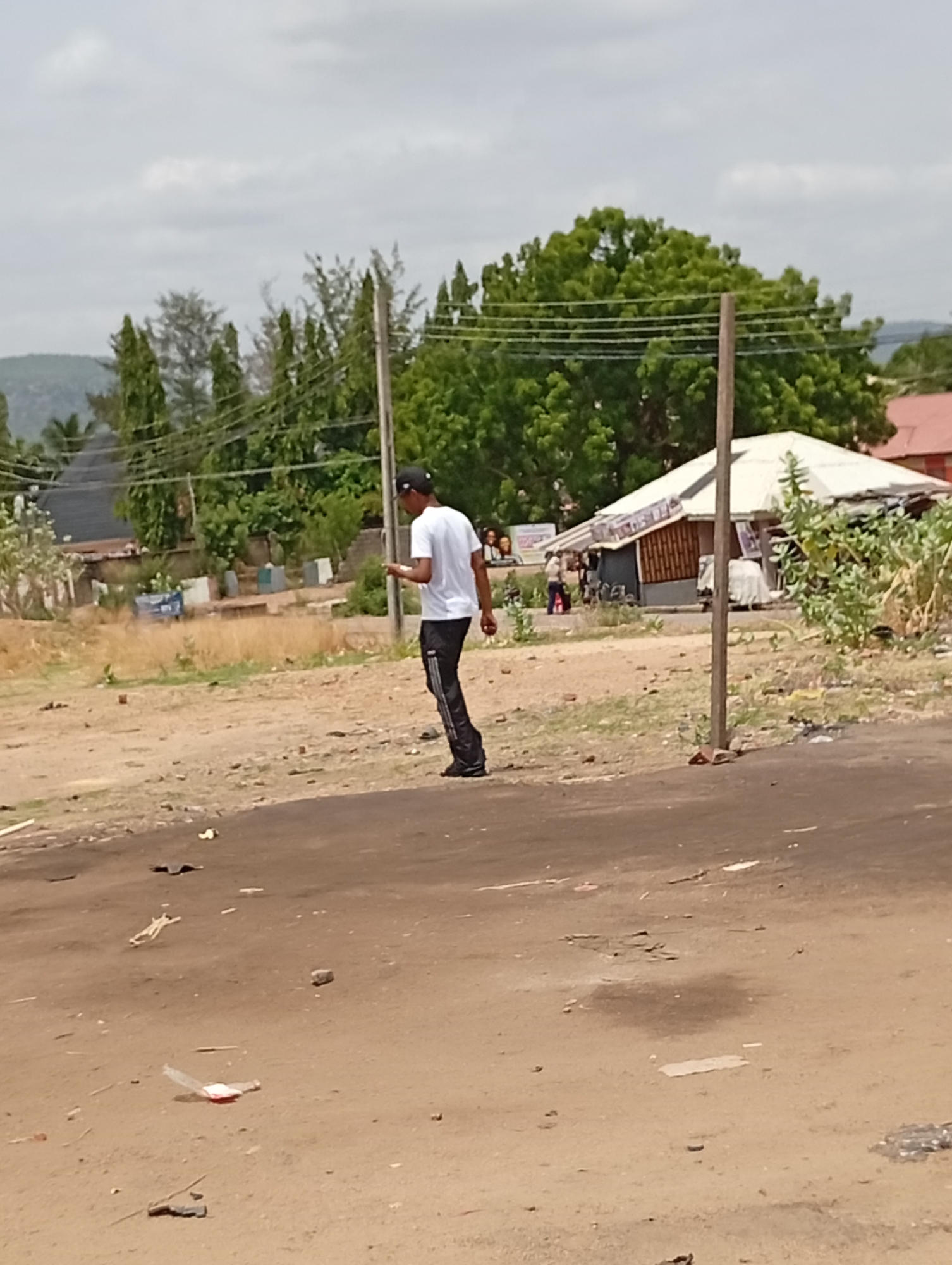 The image depicts a scene in Lokoja, Nigeria. A young man wearing a white t-shirt, black pants, and a dark cap is walking on a dirt road, appearing to look at something in his hand, possibly a phone. The road is cracked and strewn with litter. In the background, there's a building with a white roof and exposed wooden support beams, suggesting a small shop or informal business. Several people are visible near the building. Further back, there are green trees, utility poles, and a distant hill. The sky is overcast.