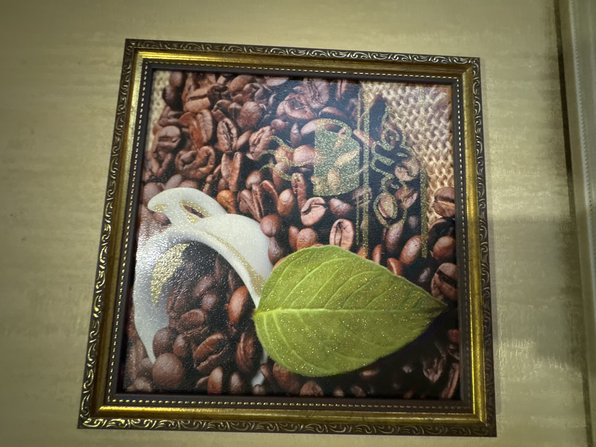 A framed artwork is displayed on a textured, pale gold wall. The artwork is rectangular and features a dark brown matting around a central image. The image depicts a close-up of roasted coffee beans, with a white coffee cup and saucer partially visible at the top. A bright green, glitter-covered leaf is positioned prominently in the foreground, overlapping the coffee beans. The frame itself is ornate, with a dark brown inner layer and a decorative gold outer layer featuring intricate scrolling patterns. The wall's texture is consistent, with subtle horizontal lines and a slightly mottled appearance. The lighting appears to be soft, casting gentle shadows. There is no indication of a specific location within Poltava, Ukraine, as the scene is confined to the artwork and the wall it is mounted on. No people or actions are depicted.