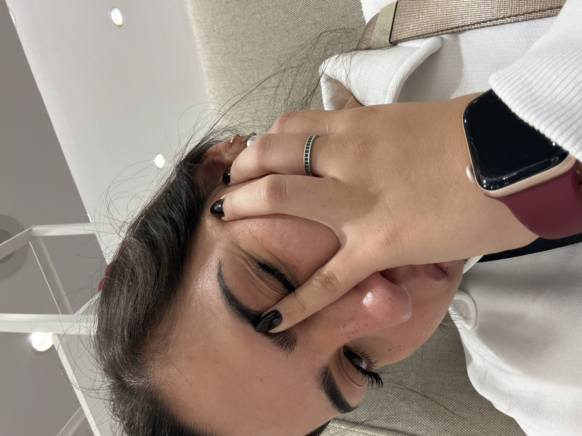A close-up, angled shot shows a person obscuring their face with their hand. The person has dark hair and wears a silver ring with small, dark stones on their ring finger. The fingernails are painted black. A smartwatch with a black face and a maroon band is visible on their wrist. They wear a white shirt with a visible tan strap, possibly part of a bag or seatbelt. The background includes a beige textured surface, likely a couch or other upholstered furniture, and a white ceiling with recessed lighting. The location is likely indoors. There is no definitive information available to specifically place this photo in San Antonio, United States of America.