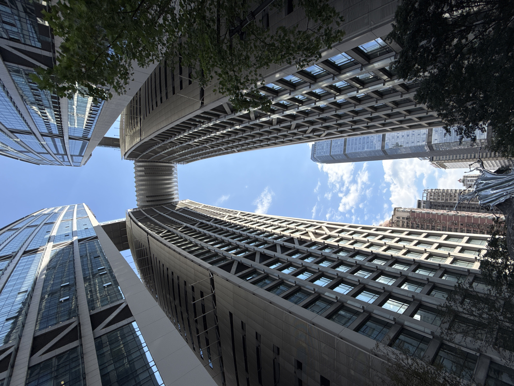 The image captures a low-angle view of a modern architectural complex in the Yuzhong District of China. Two towering, curved skyscrapers dominate the foreground, connected at a high elevation by a distinctive, segmented bridge structure. The facades of these buildings are clad in glass and metal, reflecting the bright blue sky and scattered white clouds. Additional skyscrapers, varying in height and design, are visible in the background, contributing to a dense urban landscape. Lush green trees are interspersed throughout the scene, some framing the buildings and adding a touch of nature to the concrete jungle. The lighting suggests it is daytime, with sunlight casting shadows and highlighting the architectural details.