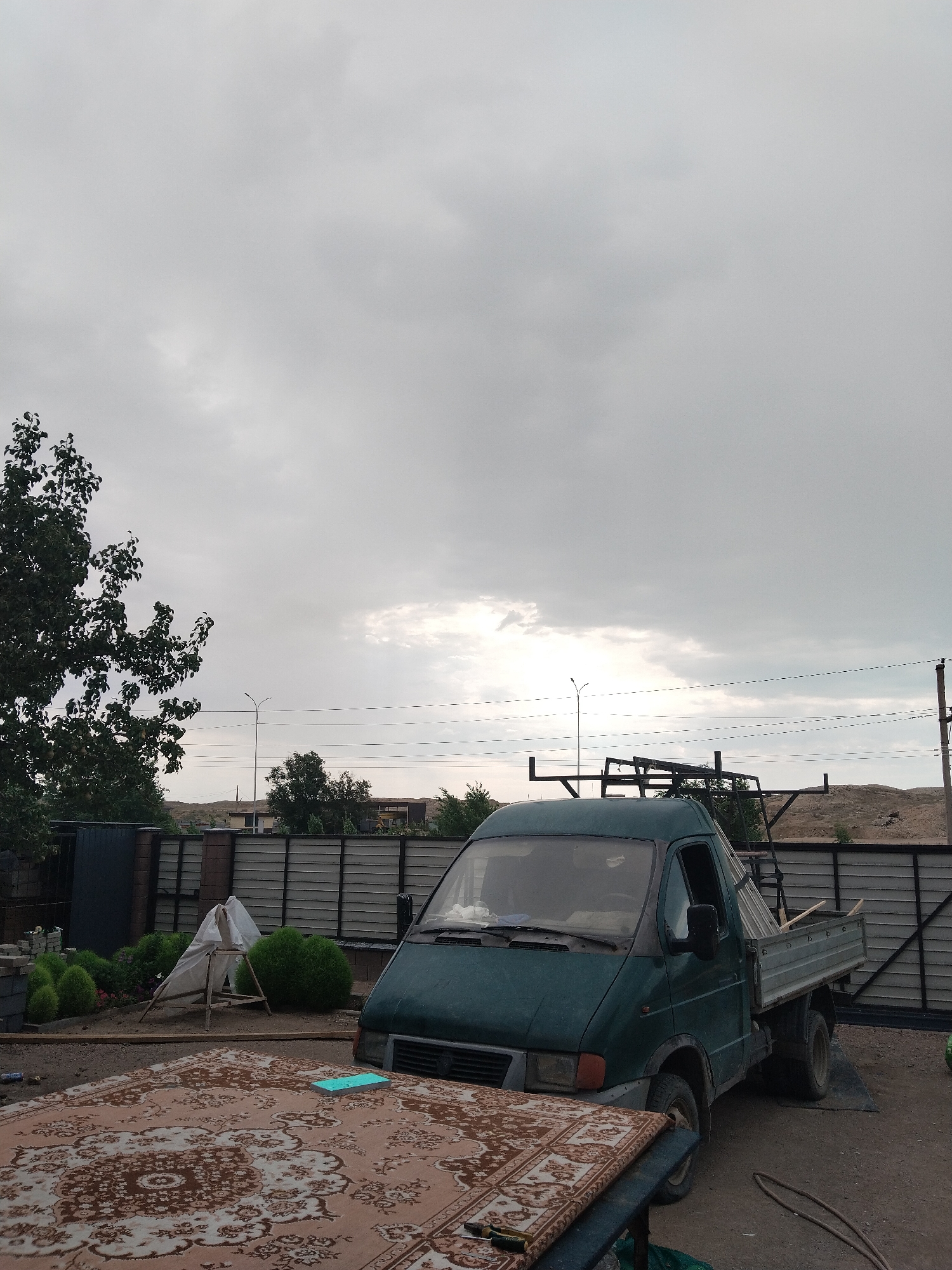 The image captures a scene in the city of Balqaş, Kazakhstan, under an overcast sky. In the foreground, a portion of a patterned rug is visible, placed on a surface. To the right, a dark green utility truck is parked, with its bed loaded with materials. Behind the truck, a grey metal fence runs horizontally across the frame, separating the immediate area from a backdrop of a dusty, arid landscape with some sparse vegetation and structures. To the left of the truck, there's a small construction area with a tarp-covered wooden structure and some round bushes. A tall tree with dense foliage stands in the far left of the image. Overhead power lines are strung across the sky, supported by poles. The overall impression is one of a semi-urban or peri-urban environment.