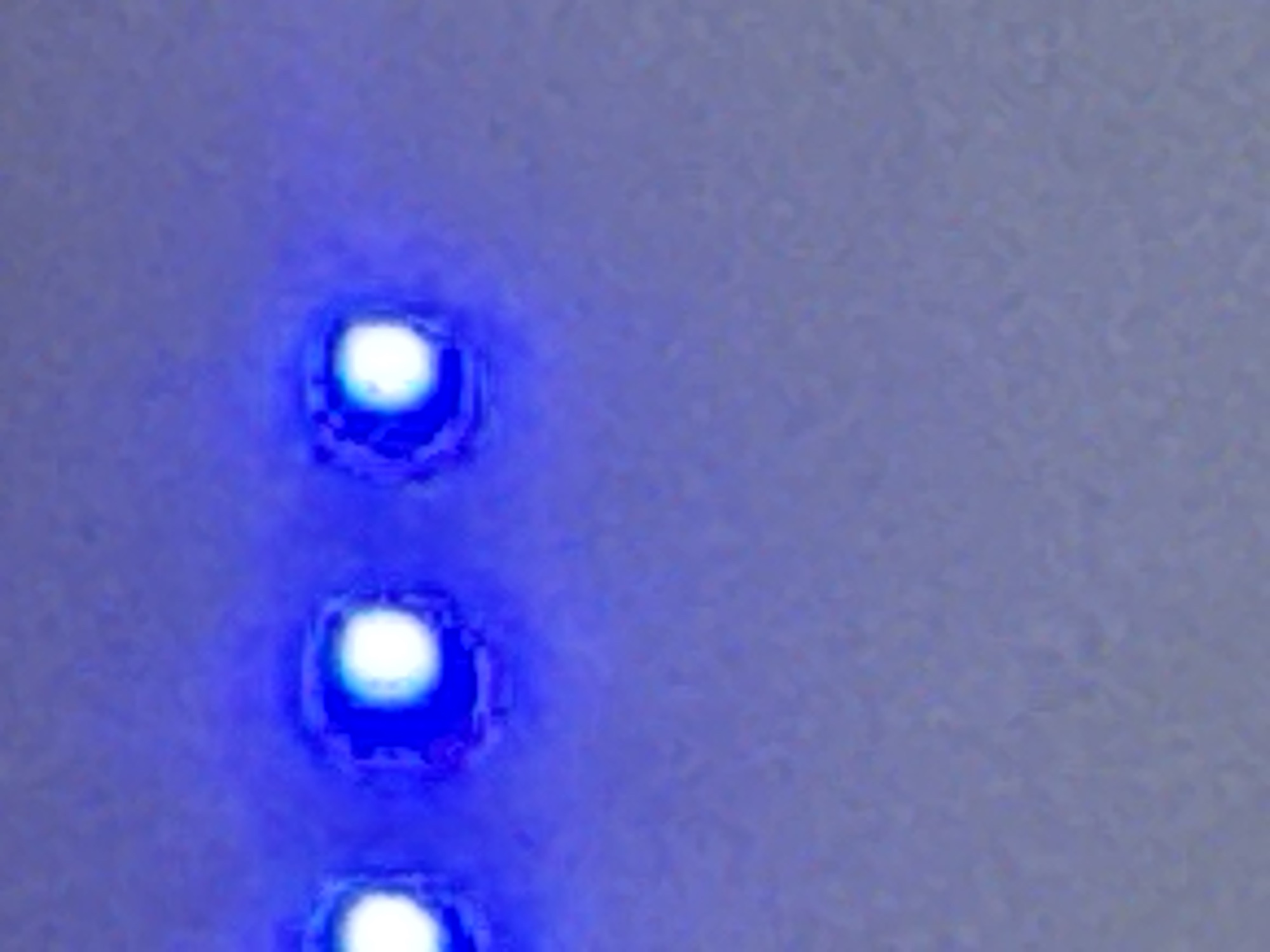 The image is a close-up, blurry, and poorly lit photograph of three illuminated circular blue lights arranged horizontally. The background is a textured, light-colored surface, possibly a wall or electronic device casing, with a subtle purple hue. No people, animals, or recognizable landmarks are present. The image does not depict any elements that would allow for the identification of its location in Navbahor, Uzbekistan.