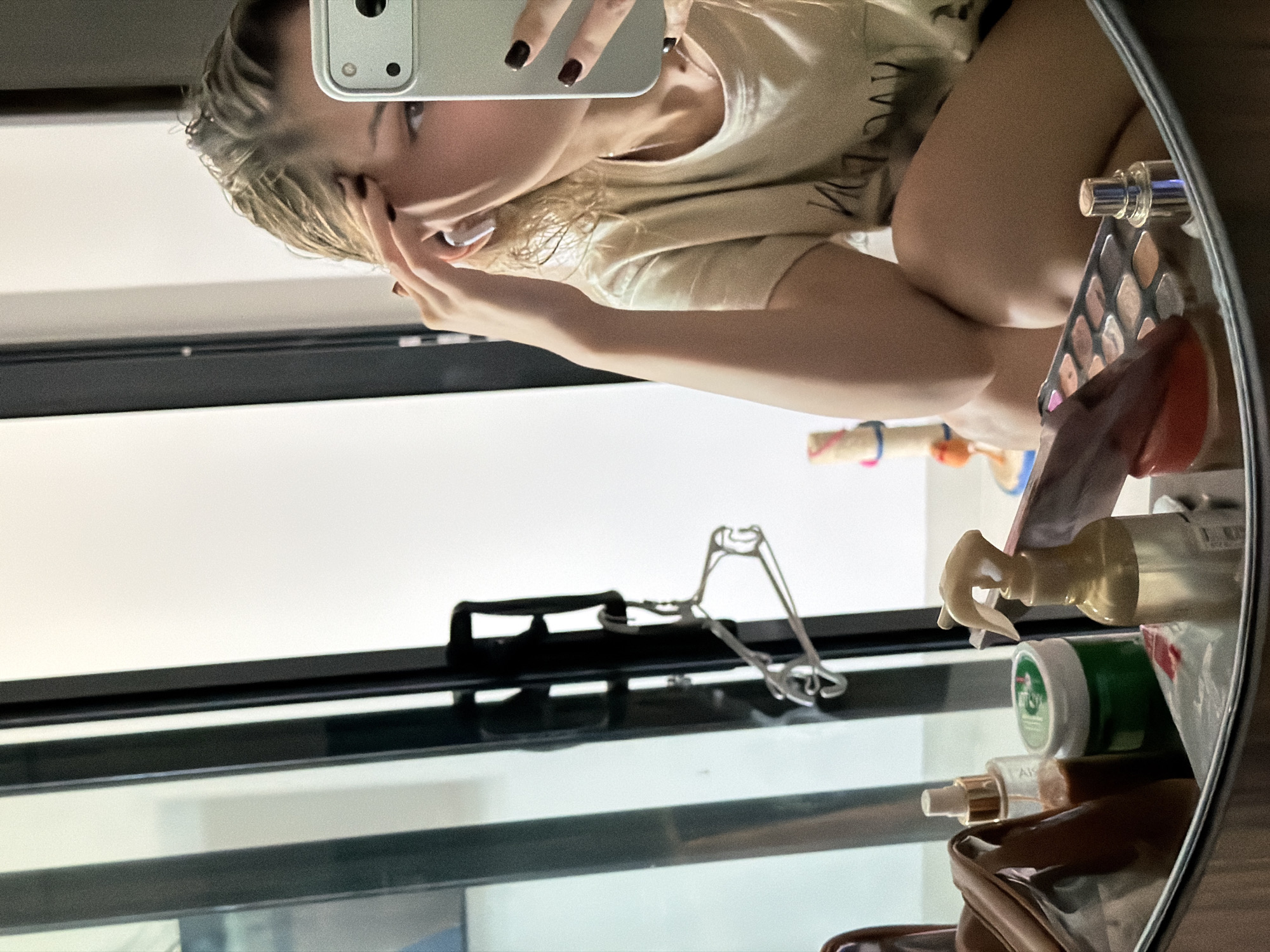 A young Caucasian woman with wet blonde hair and dark nail polish takes a mirror selfie. She is wearing a light-colored t-shirt with an unreadable dark graphic on it. She has one hand touching her forehead and the other holding a silver smartphone, which is capturing the reflection. She is wearing white earbuds. In the foreground, a vanity mirror reflects a collection of makeup items, including an eyeshadow palette, a small silver spray bottle, a green jar, and a brown cosmetic bag. Behind her, a tall, dark-framed doorway or partition is visible, with a metal coat hanger hanging from it. The background is predominantly white. The scene appears to be indoors, possibly a bathroom or dressing area. There is no clear indication of the location within Đà Nẵng, Vietnam.
