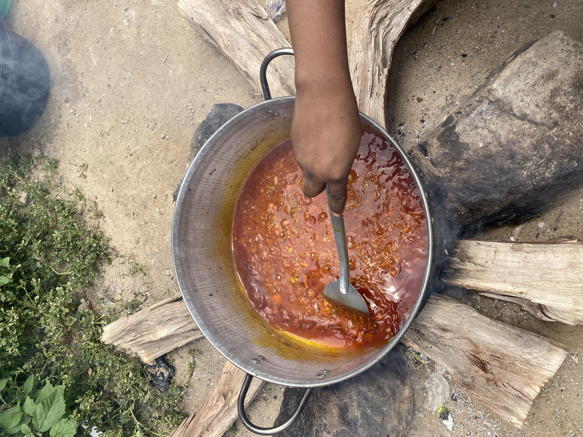 An overhead view captures a dark-skinned arm and hand stirring a bubbling, reddish-orange stew within a large, metallic pot using a silver spatula. The pot is positioned outdoors on light brown sandy ground, supported by several weathered wooden logs and a large dark rock. Wisps of smoke emanate from beneath the pot, indicating an active open fire. The stew itself is thick, showing small solid components and a visible layer of oil on its surface. The ground surrounding the cooking area contains scattered small pebbles and debris, including crumpled plastic bags and white fragments. Green vegetation, consisting of small-leafed plants and larger broad leaves, is visible along the upper left and right periphery of the frame. A dark, cylindrical container emitting smoke is partially visible in the upper right background. This scene is located in the city of Kubwa, Nigeria.