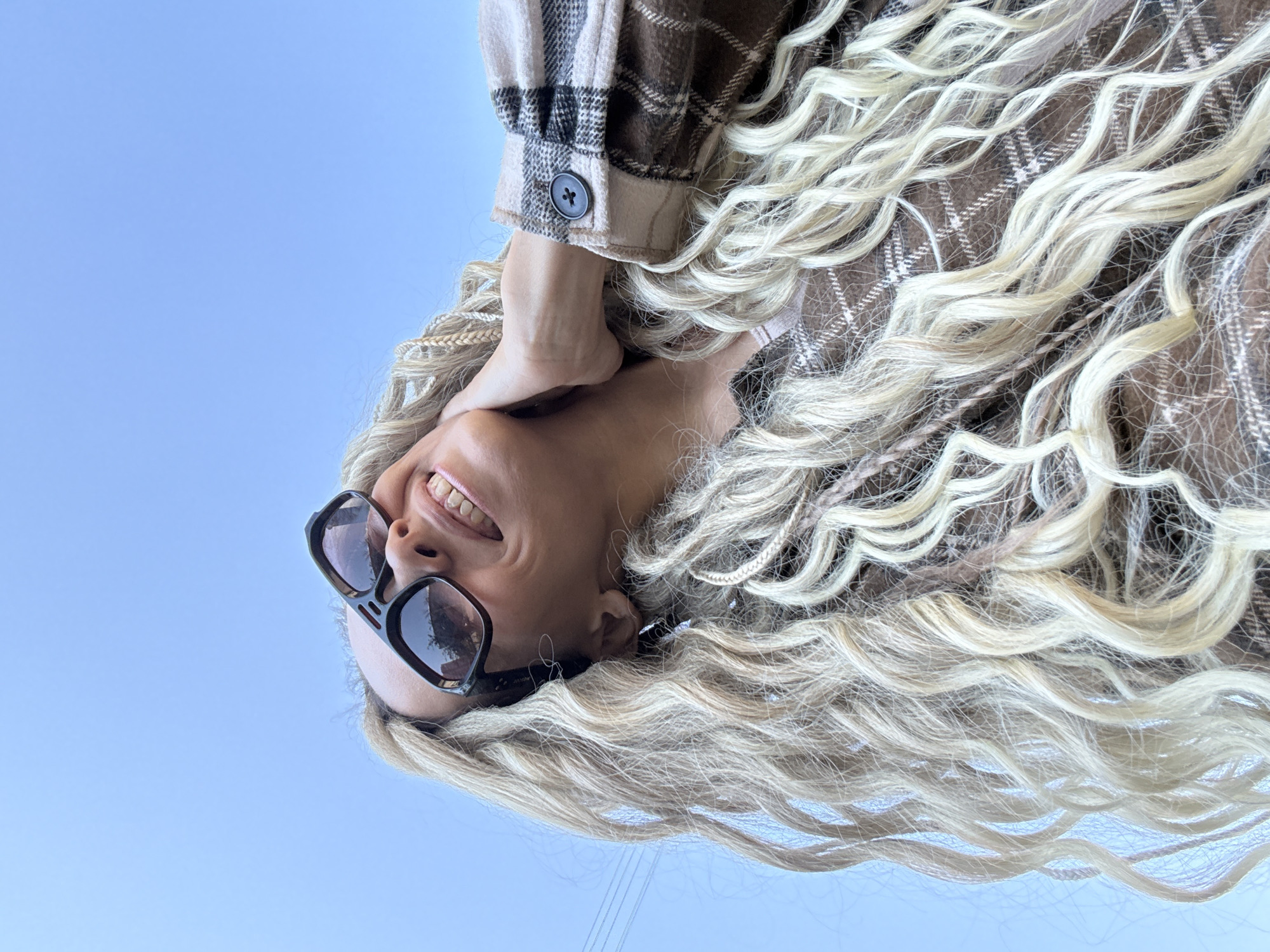 An upside-down eye-level view of a woman wearing sunglasses and a brown plaid shirt, taken outdoors under a clear, light blue sky. Her blond hair is styled with a mix of braids and loose waves. Her right hand is touching her neck. The reflection in her sunglasses includes a leafy tree. Based on the electrical lines in the background and the presence of a similar clear blue sky, the location may be in Rusanovo, Russia, but there's no definite confirmation within the image itself.