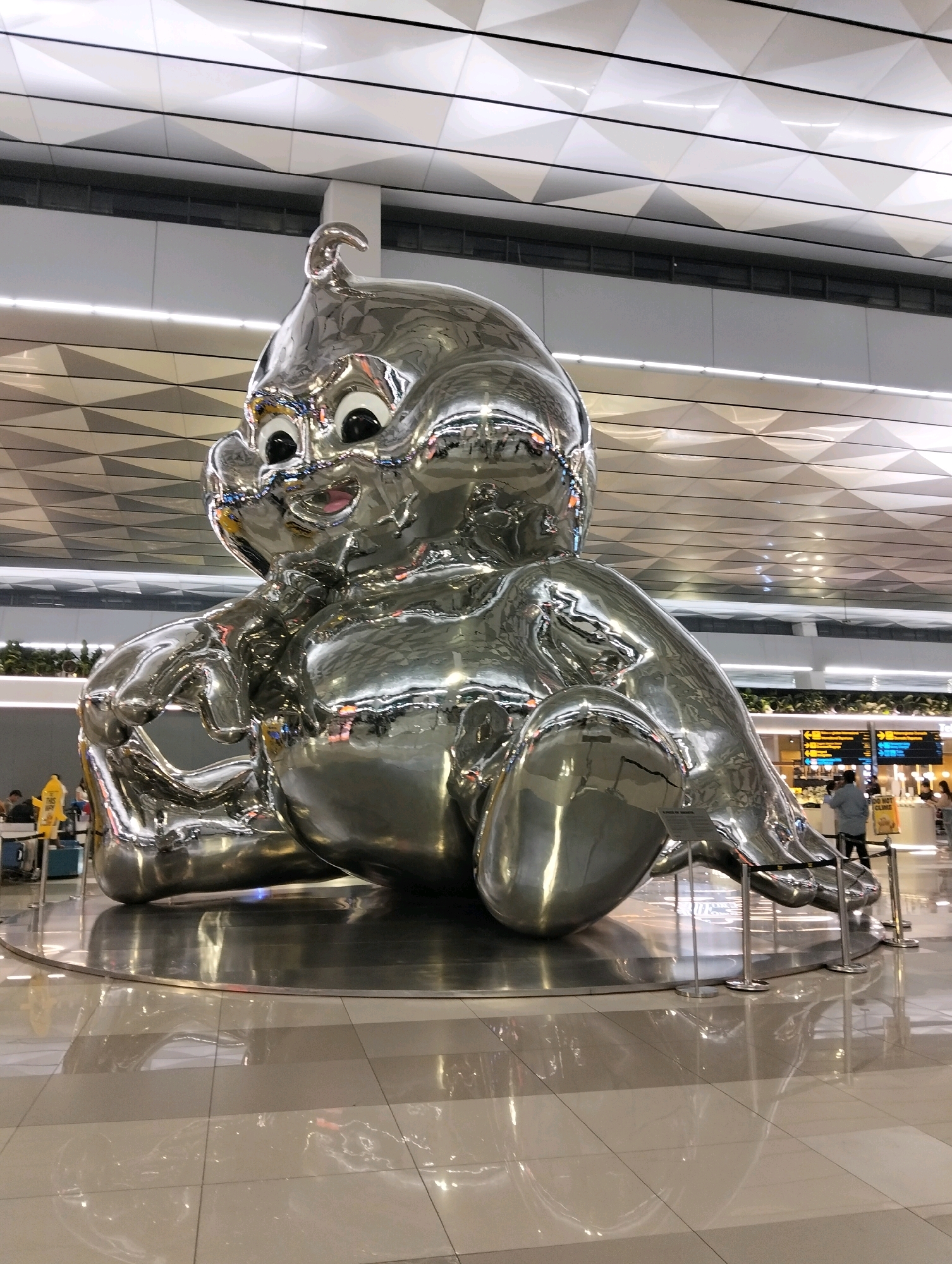A large, shiny, silver sculpture of a baby with large eyes, a smile, and a small tuft of hair on its head is the central subject of the image. The sculpture is positioned in the middle of a bright, modern interior space with a tiled floor that reflects the sculpture. Above, the ceiling is a complex geometric design with recessed lighting. In the background, there are elements of an airport terminal, including signs with arrows, a yellow information stand with text, people walking, and baggage carts. The overall environment suggests a public space, possibly an airport or a large shopping mall in Pajang, Indonesia.