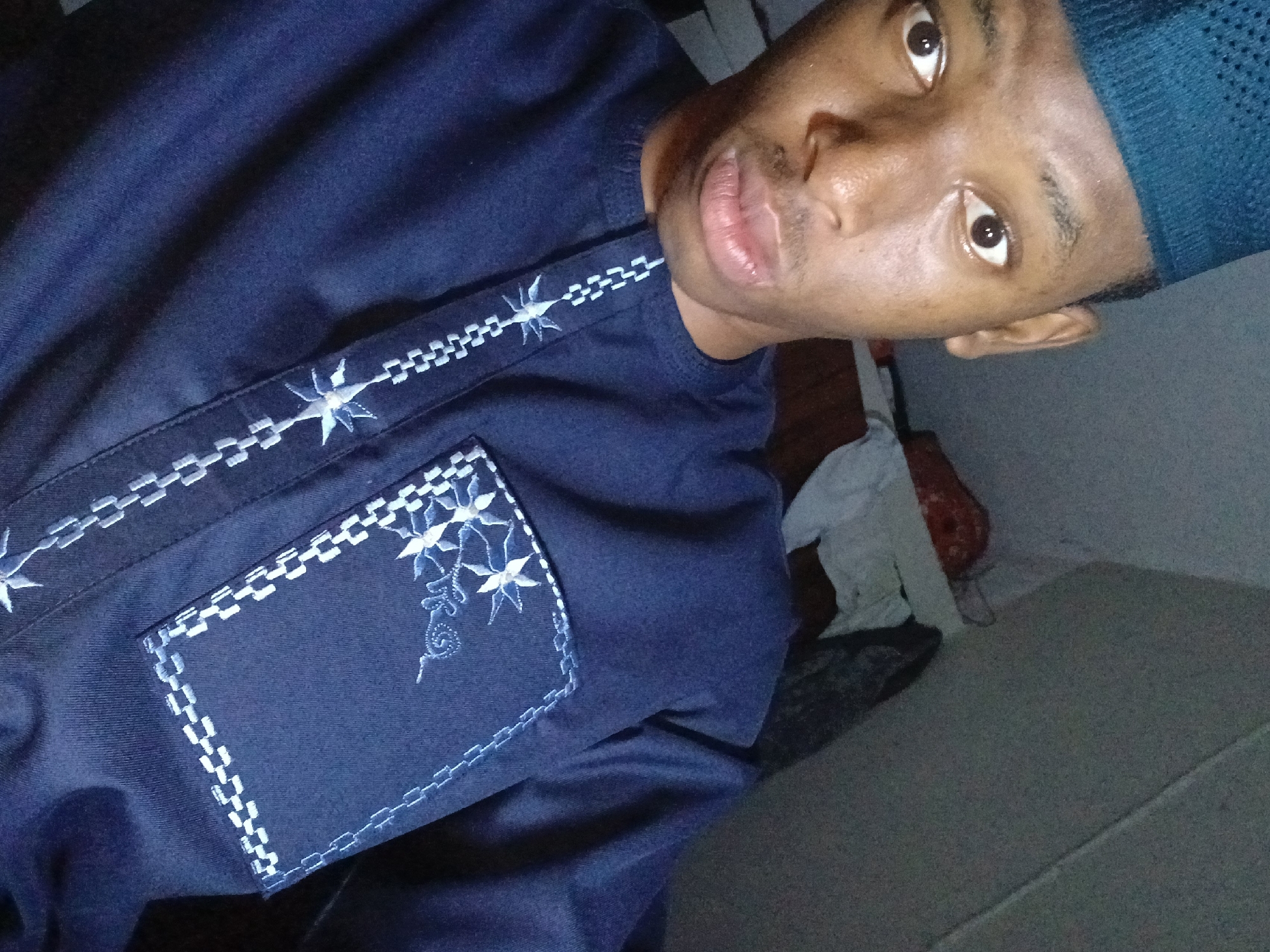 A young man, likely in his late teens or early twenties, is pictured in a selfie. He has dark skin, brown eyes, and short hair, covered with a patterned blue cap. He wears a dark blue shirt or tunic with white embroidery around the collar and pocket. The photo is taken indoors, with glimpses of a wall and a partially visible object that appears to be bedding in the background. The location is not discernible as Lagos, Nigeria based on the visual information.