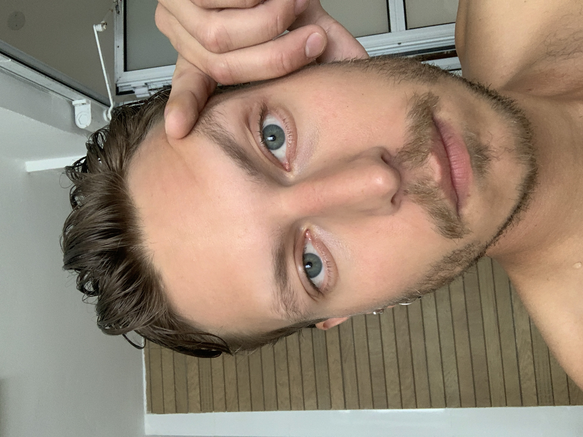 A close-up, rotated image shows a light-skinned man with light eyes, a beard, and wet, dark hair. His fingers are touching his forehead. He appears to be in a bathroom or shower area. The backdrop includes light-colored vertical slats or paneling and white tilework. The location in Tel-Aviv, Israel, is not directly evident from the image.