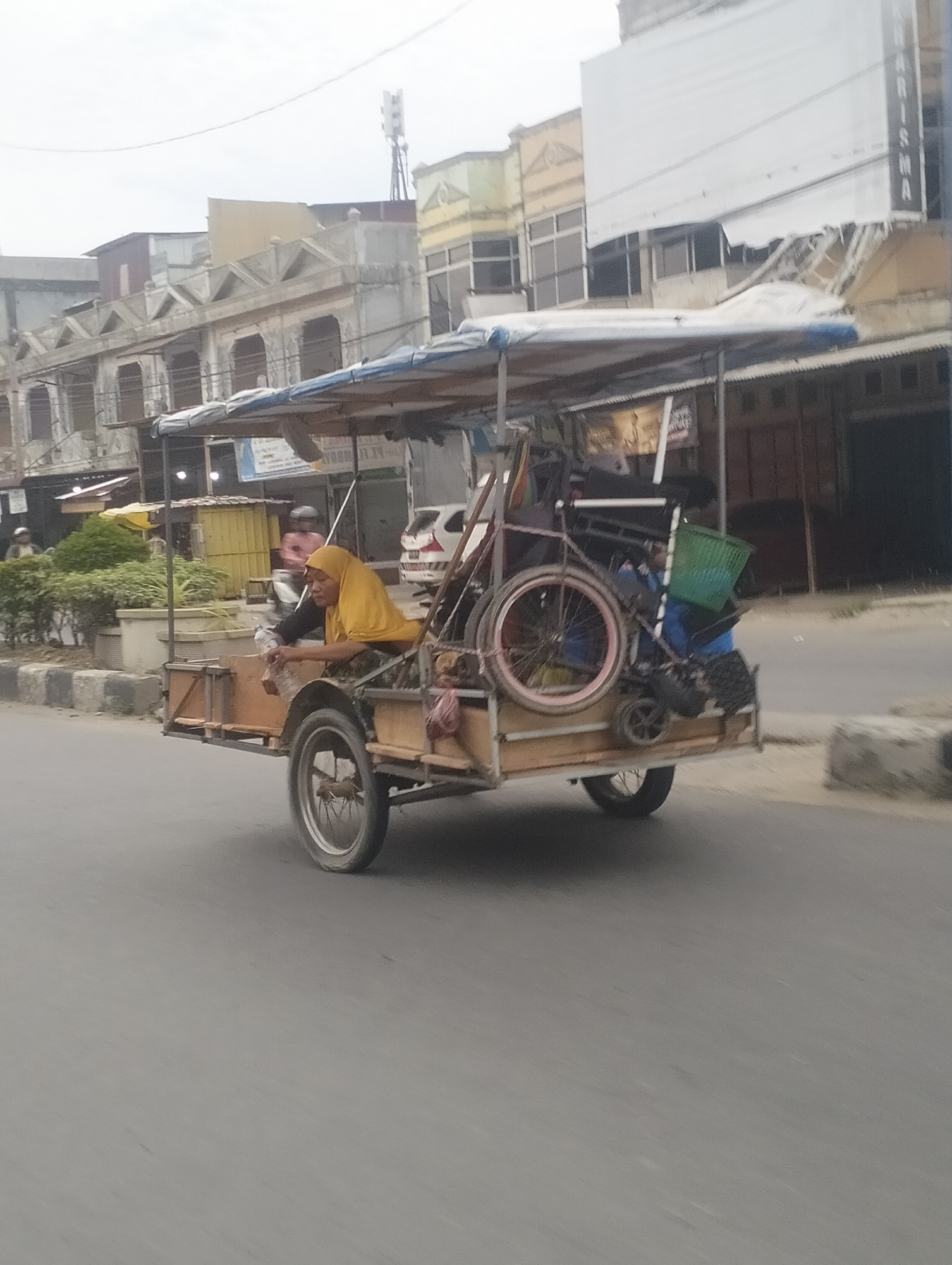 In Lhokseumawe, Indonesia, on a cloudy day, a pedicab driver wearing a yellow hijab is operating a heavily loaded pedicab. The pedicab is laden with various items, including what appears to be a bicycle, a green basket, and other miscellaneous objects. The driver is seated in the pedicab, seemingly in motion, with their hands on the controls. The pedicab is traveling on a paved road. In the background, buildings line the street, some appearing to be shops or businesses with signage. The overall scene depicts a mode of transportation commonly used in urban areas in Indonesia for both passengers and cargo.