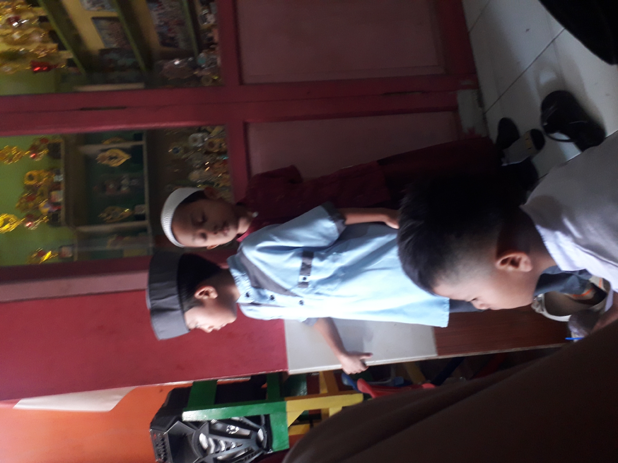 The image depicts three boys in what appears to be an indoor setting, possibly a classroom or a community hall in Cankuang, Indonesia. The boy in the foreground on the left is wearing a grey peci (cap) and a light blue tunic with grey trim. He is facing to the right, with his left arm resting on a white surface. To his right stands another boy wearing a white peci and a dark red tunic. He is also looking to the right. In the background, a third boy is partially visible, wearing a white shirt and facing left. Behind the boys, shelves are filled with trophies, suggesting an award ceremony or a place where achievements are displayed. The room is painted in red and has a window or door with red trim. The floor is tiled. The overall atmosphere seems casual and communal.