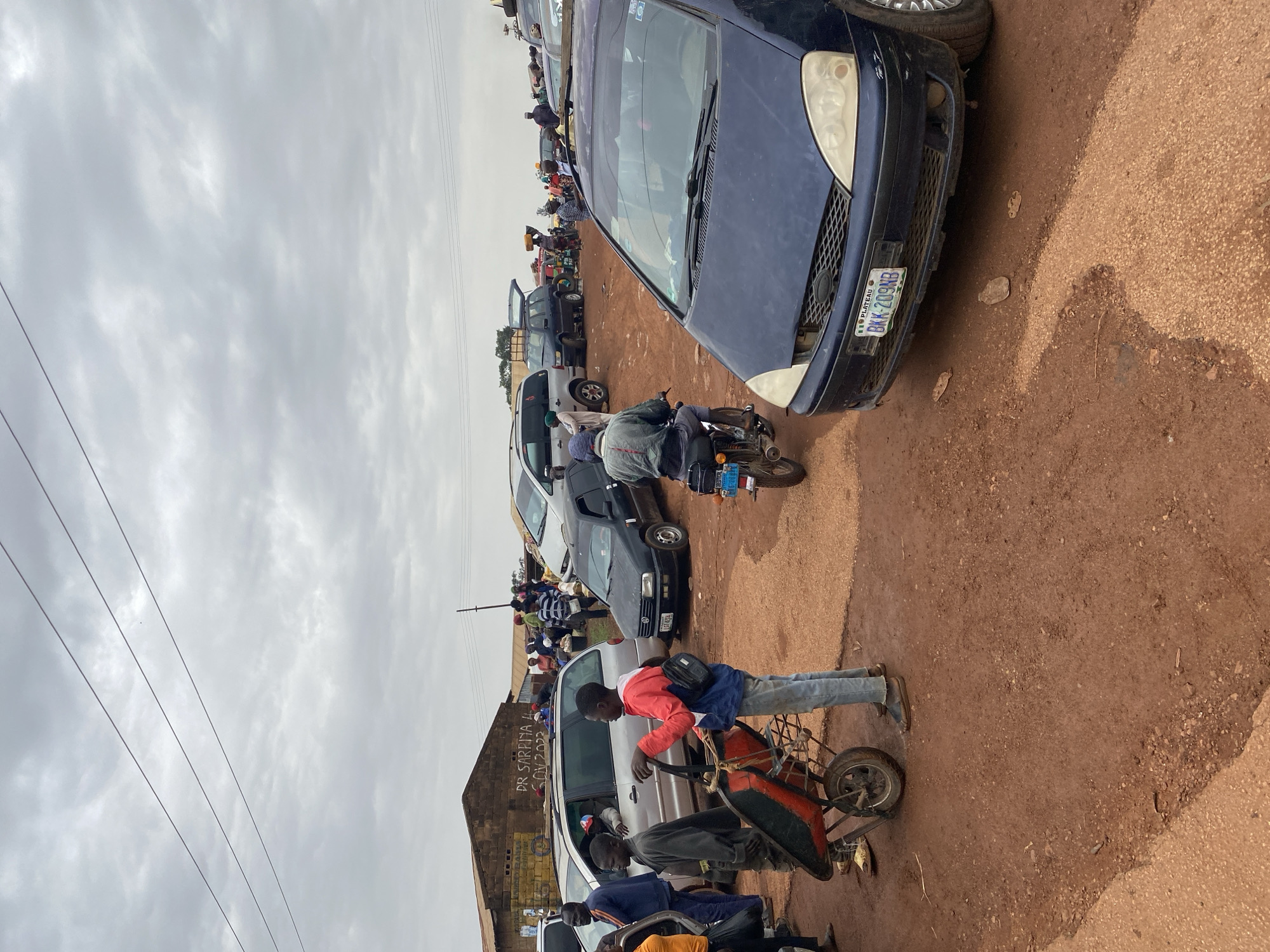 The image depicts a congested street scene in Durumi, Nigeria. Several vehicles, including cars and motorcycles, are stalled in a traffic jam on a red dirt road. A group of people are milling around the vehicles. One individual is pushing a wheelbarrow with what appear to be items being transported. A building is visible in the background, marked with signage. The sky is overcast, indicating cloudy weather. The license plate on a blue car reads "BKX-209NB".