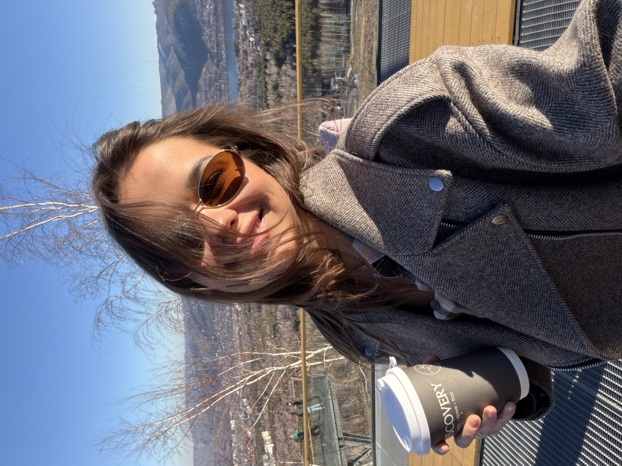A young woman with shoulder-length brown hair, wearing sunglasses and a gray herringbone jacket, holds a gray-and-white paper coffee cup labeled "Discovery" in Russian and English. She is smiling at the camera. Behind her is a cityscape including a river, trees, mountains, and blue sky. A metal grate is visible below and a wood railing surrounds the area. This is likely a selfie taken from the observation deck of the Torgashinsky Ridge in Krasnoyarsk, Russia.
