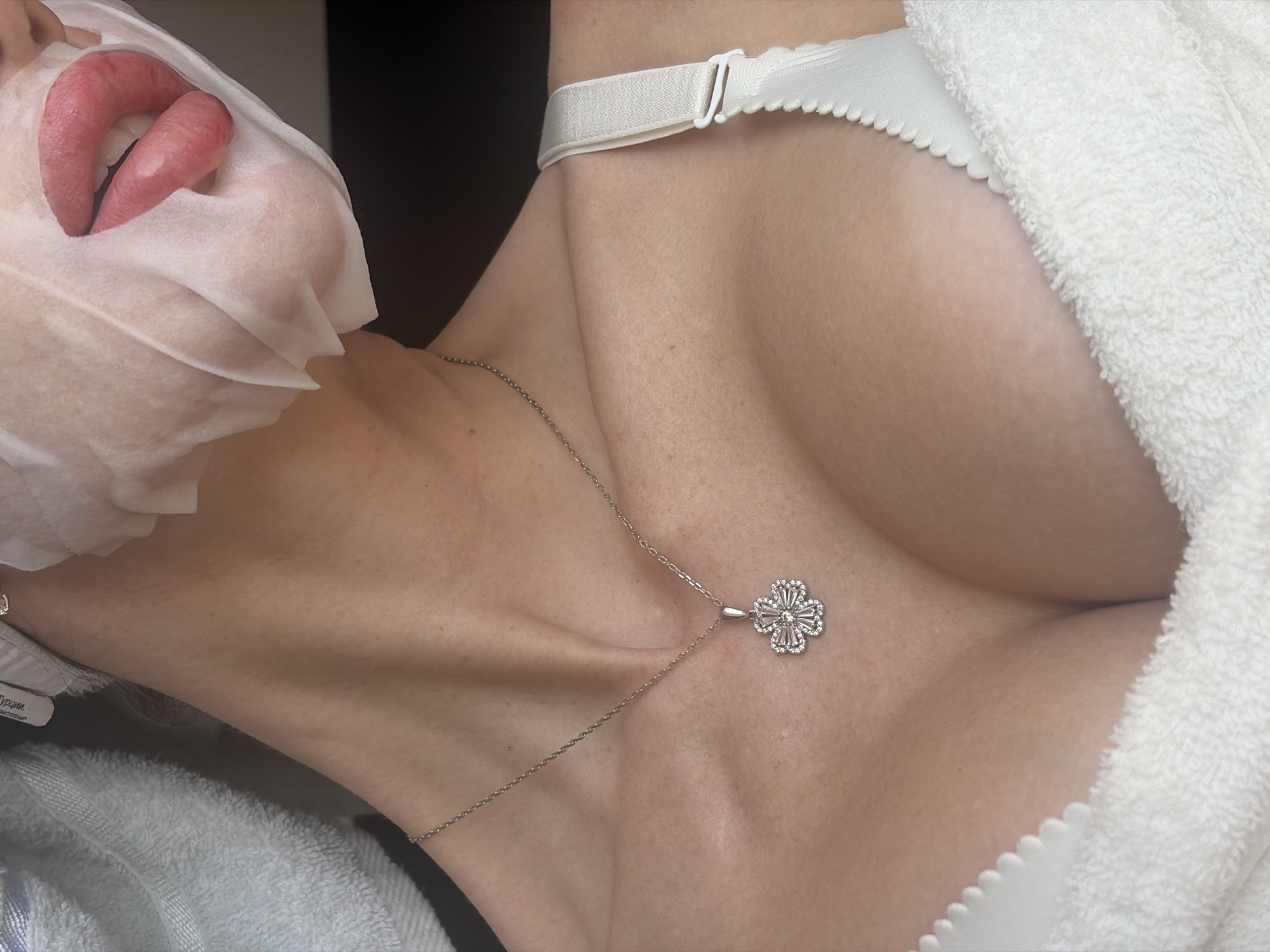 An extreme close-up shot shows a person with a facial mask covering most of their face except their lips, which are parted. The person wears a white bra and a delicate silver necklace with a clover-shaped pendant. A white towel is draped around their shoulders and chest. The image does not contain enough information to determine its location in Saint Petersburg, Russia.