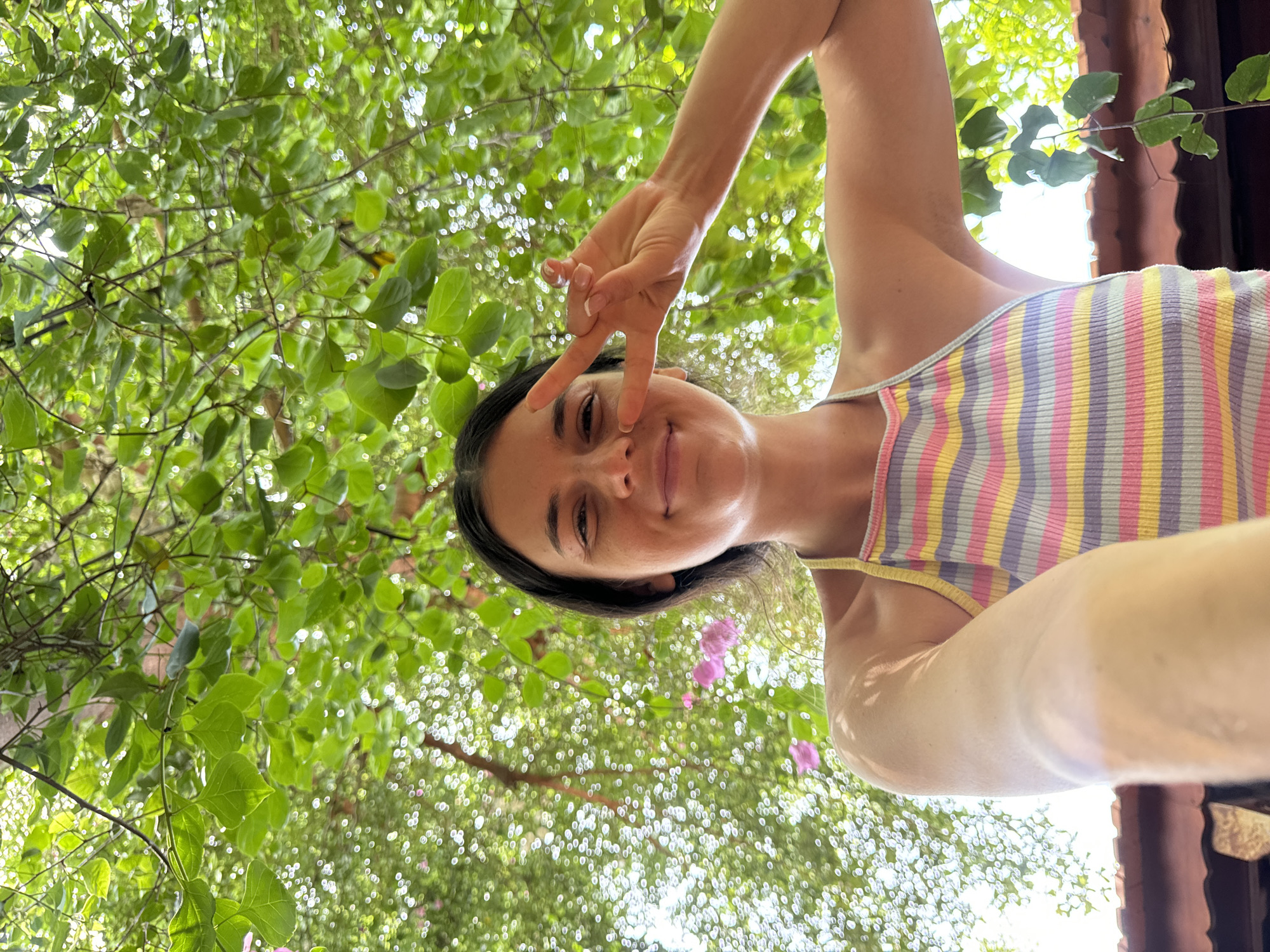 A young woman, with long dark hair, is taking a selfie outdoors under a canopy of green leaves. She is smiling and holding up two fingers in a peace sign. She is wearing a multicolored striped halter top. The background is blurred, showing more green foliage and hints of pink flowers. Below her, a tiled roof is visible. The scene appears to be in a tropical environment, possibly a garden or a resort in Mũi Né, Vietnam.