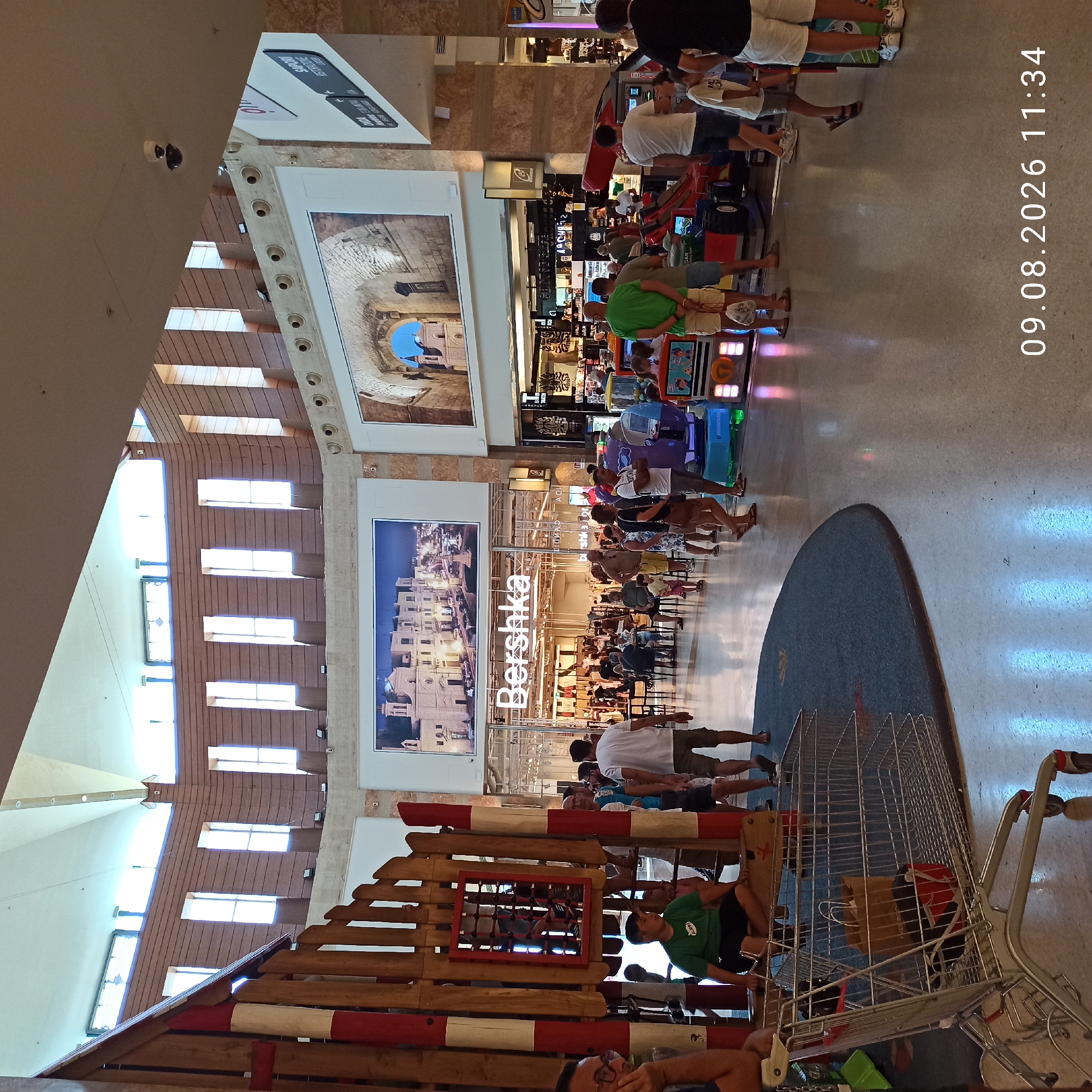 The image captures an indoor scene, likely a shopping mall, with architectural features suggesting a modern, open-plan design. A large, multi-story structure with wooden paneling and arched window openings dominates the background. The ceiling is high and appears to be a white, vaulted structure with visible lighting.
In the foreground, a wooden playground structure is visible on the left, featuring red and white striped poles. A shopping cart with a brown paper bag and a red item inside is positioned in front of the playground.
A central element of the scene is the "Bershka" clothing store, identifiable by its prominent signage above a storefront with mannequins and clothing displays. Adjacent to Bershka, to the right, are other retail establishments and gaming machines, including a purple and a red racing-themed arcade game with children around them.
Several people are present, scattered throughout the mall. Some are walking, while others are standing or sitting. A young boy in a green t-shirt and black shorts is seated on a wooden beam of the playground structure. An adult man in glasses is seated on the left edge of the frame. Various other individuals, including families and couples, are visible browsing, playing, or walking.
The floor is a polished, reflective surface, likely concrete or stone, with light patterns reflecting from the overhead lighting and possibly from the arcade machines. The overall ambiance suggests a daytime scene in a busy commercial environment.
The timestamp "09.08.2026 11:34" is overlaid on the bottom right of the image, indicating the date and time the photograph was taken.
This scene is located within the **Marecity Shopping Centre** in the city of Molfetta, Italy.