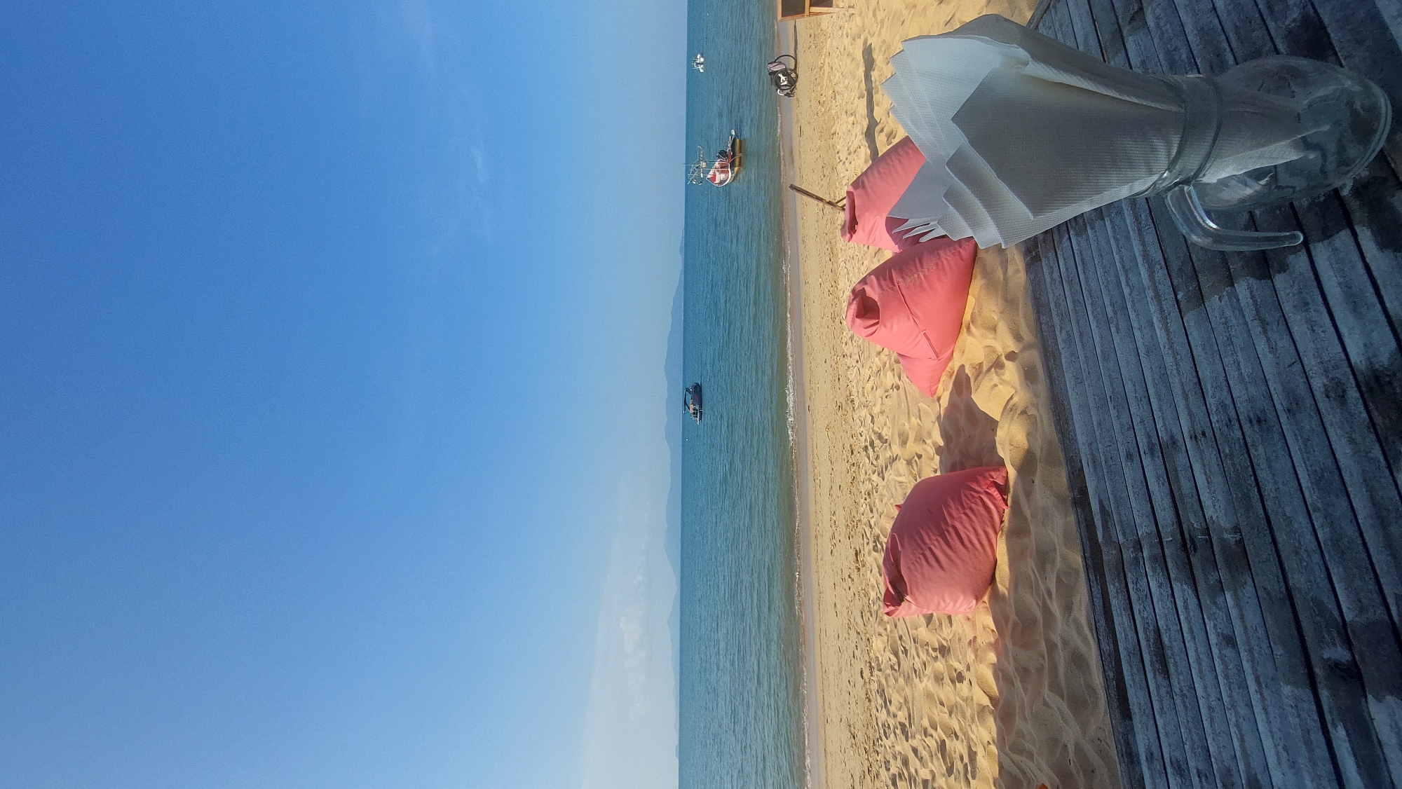 This image depicts a sunny and serene beach scene, likely captured at Baan Mae Nam, Thailand, given the location context. The shot is vertically oriented (or rotated), presenting a bright blue sky on the left, merging into the tranquil blue-green ocean and a distant, hazy landmass.
Several small boats dot the calm water; one closer to the shore is white and yellow with individuals visible, while another darker boat floats further out. A stretch of light-colored sandy beach runs along the coastline, where several vibrant pink bean bag cushions are scattered, suggesting a casual lounging area. These bean bags cast clear, elongated shadows, indicating bright sunlight.
To the right, a wooden deck, composed of weathered planks, extends into the foreground. On this deck, a clear glass pitcher is prominently placed, containing neatly folded white napkins or paper towels secured by a translucent band. The pitcher also casts a distinct shadow.
The scene conveys a peaceful atmosphere, characteristic of a tropical vacation spot. The clear sky, calm water, and bright sunshine suggest good weather, likely during the mid-morning or afternoon. No visible text is present in the image.