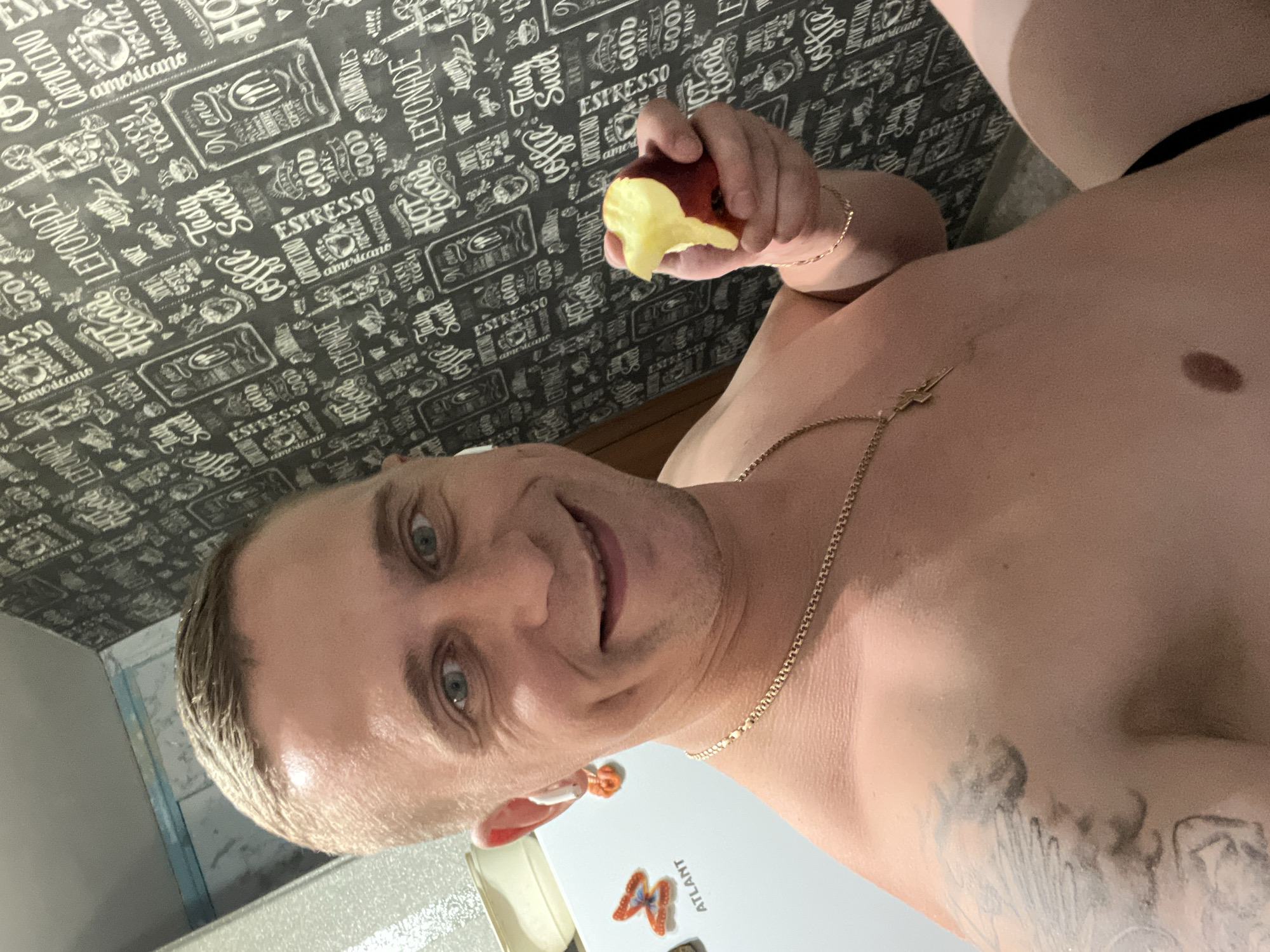 A shirtless man is taking a selfie in what appears to be a kitchen. He has fair skin, short blonde hair, and blue eyes. He is smiling and holding a piece of fruit in his right hand. He is wearing a gold chain around his neck with a lightning bolt pendant, and white wireless earbuds in his ears. On the wall behind him is a black and white patterned wallpaper with coffee-themed designs. To his left, a white refrigerator is visible with a butterfly-shaped magnet and the word "ATLANT" printed on it. The man's left arm has a tattoo. The overall scene is consistent with an indoor residential setting. The presence of the "ATLANT" brand suggests a possible location within Minsk, Belarus, as Atlant is a major Belarusian manufacturer of household appliances.