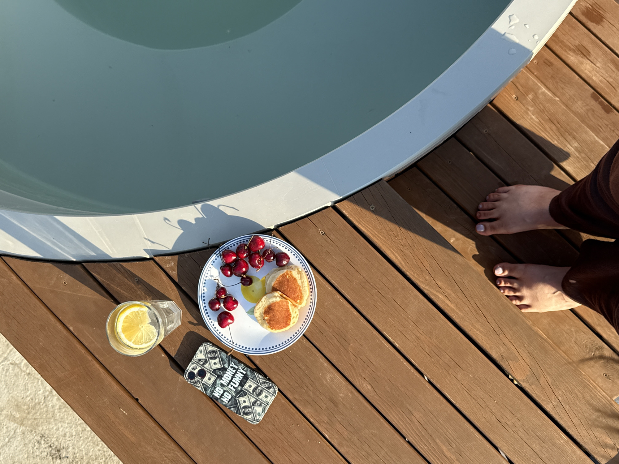 This overhead shot captures a serene moment on a wooden deck beside a pool. The pool's pale blue water fills the upper left of the frame, its white rim casting a subtle shadow. On the right side, a person's bare feet, with painted toenails, rest on the warm brown wooden planks. The person is wearing dark brown pants.
In the foreground, a white plate holds two small, golden pancakes and a cluster of bright red cherries. To the left of the plate, a clear glass contains a refreshing drink with a slice of lemon. Below the plate, a black smartphone case is visible, featuring a repeating pattern of dollar bills and the text "NO MONEY NO FUNNY".
The lighting suggests it's a sunny day, with long shadows indicating either early morning or late afternoon. The overall mood is one of relaxation and indulgence, perhaps a leisurely breakfast or snack enjoyed outdoors. The scene is set in Starokorsunskaya, Russia.