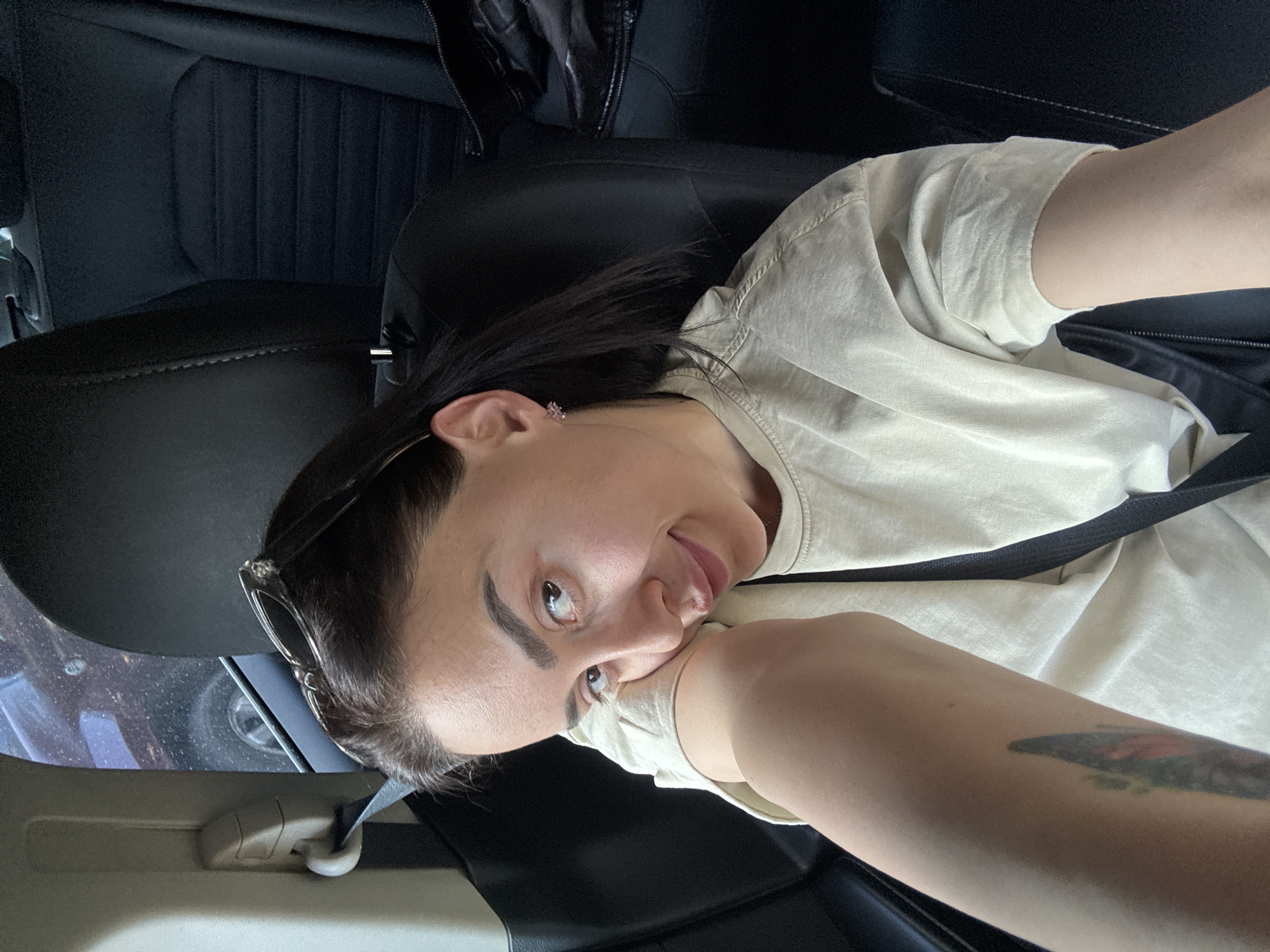 A person is captured in a selfie taken inside a car. The subject, with dark hair, prominent eyebrows, and fair skin, wears a light beige or off-white short-sleeved t-shirt. She is seated in the front passenger seat, secured by a dark seatbelt across her chest and lap. Her left hand supports her head as she looks playfully towards the camera, with a slight smile and her tongue subtly extended.
Notable details include a small, sparkly, flower-shaped earring in her left ear and dark-rimmed sunglasses perched on top of her head. A colorful tattoo, featuring shades of red, green, and black, is visible on her right forearm. The car's interior is predominantly dark, with black leather or faux leather seats. Vertical stitching is visible on the seatback. The lighting inside the vehicle appears somewhat subdued. Through the car's window, a reflection shows parts of an outdoor scene, possibly including a vehicle tire and other indistinct shapes. There is no visible text in the image.