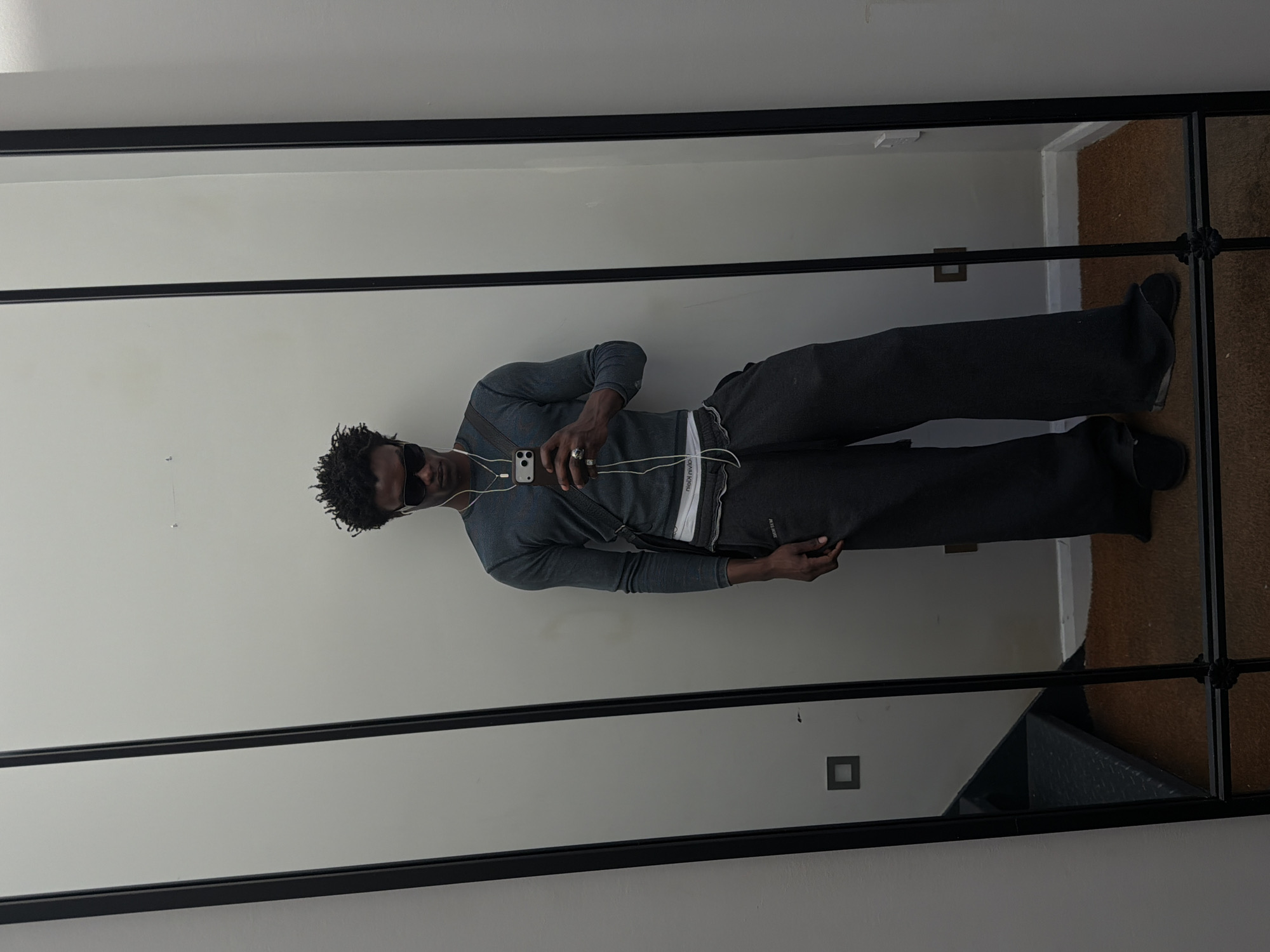 A man stands in front of a floor-length mirror with a black metal frame. The man is dark-skinned with short, curly black hair, wearing sunglasses, a grey long-sleeved shirt, grey sweatpants with a white waistband detail, and dark shoes. He is holding a smartphone in his hand and appears to be taking a photo of himself in the mirror. Earphones are connected to the phone. The mirror reflects a white or light grey wall. In the background, a portion of a brown floor covering is visible to the right. The floor appears to be a carpet. A section of the bottom right reveals a black shelf or structure. Two electrical outlet boxes are visible on the wall within the mirror’s reflection. The location is likely an interior space in London, United Kingdom.