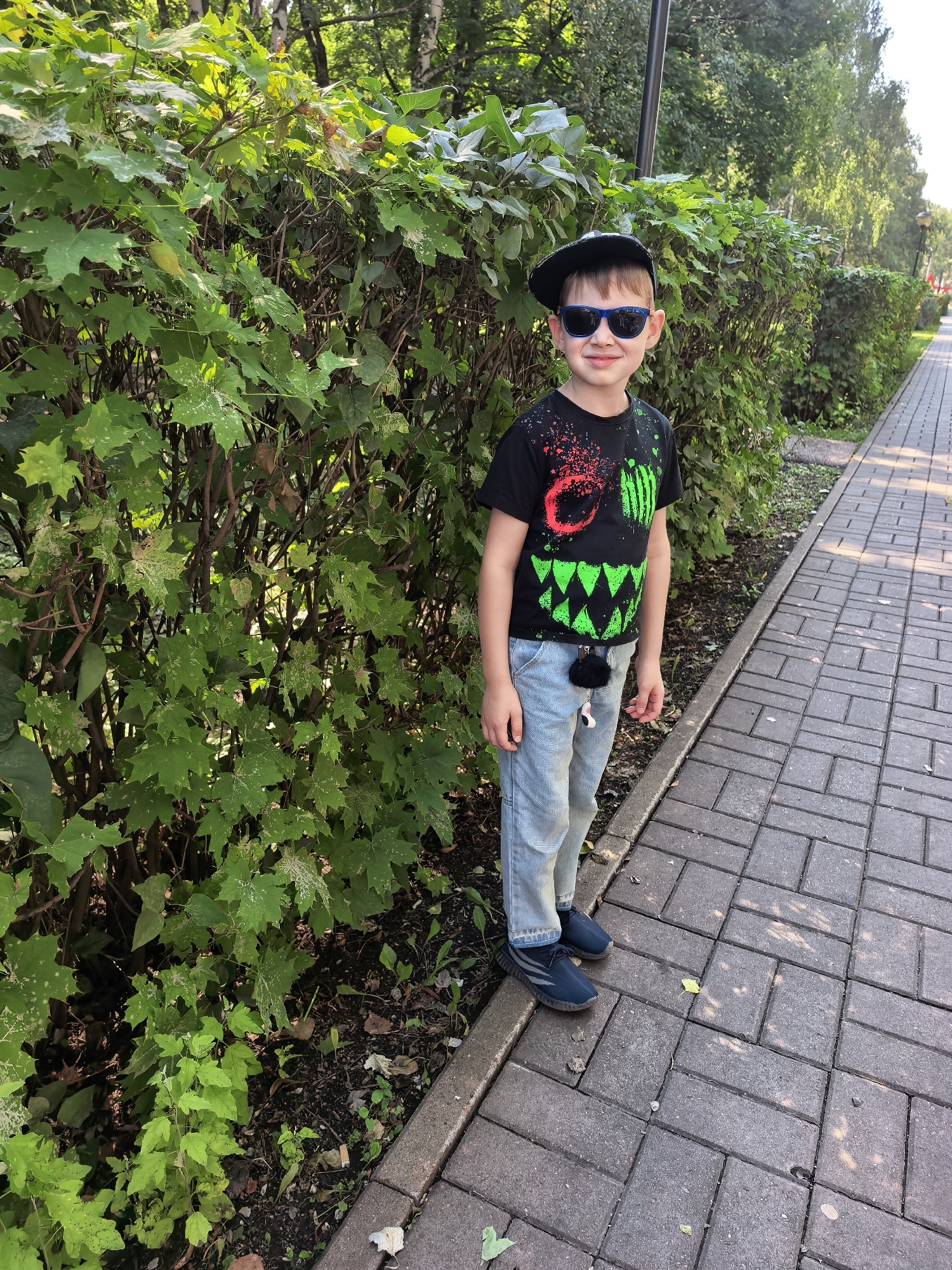 A young boy stands on a paved walkway next to a dense green hedge in Sterlitamak, Russia. He is wearing a black t-shirt with a monster-like graphic, light blue jeans, a black baseball cap, and blue sneakers. He is also wearing sunglasses. The boy is facing the camera with a slight smile. The walkway is made of rectangular grey pavers, and the hedge is made up of many leafy branches. Sunlight filters through the leaves, creating dappled patterns on the ground. In the background, trees and more foliage are visible, suggesting a park or residential area.