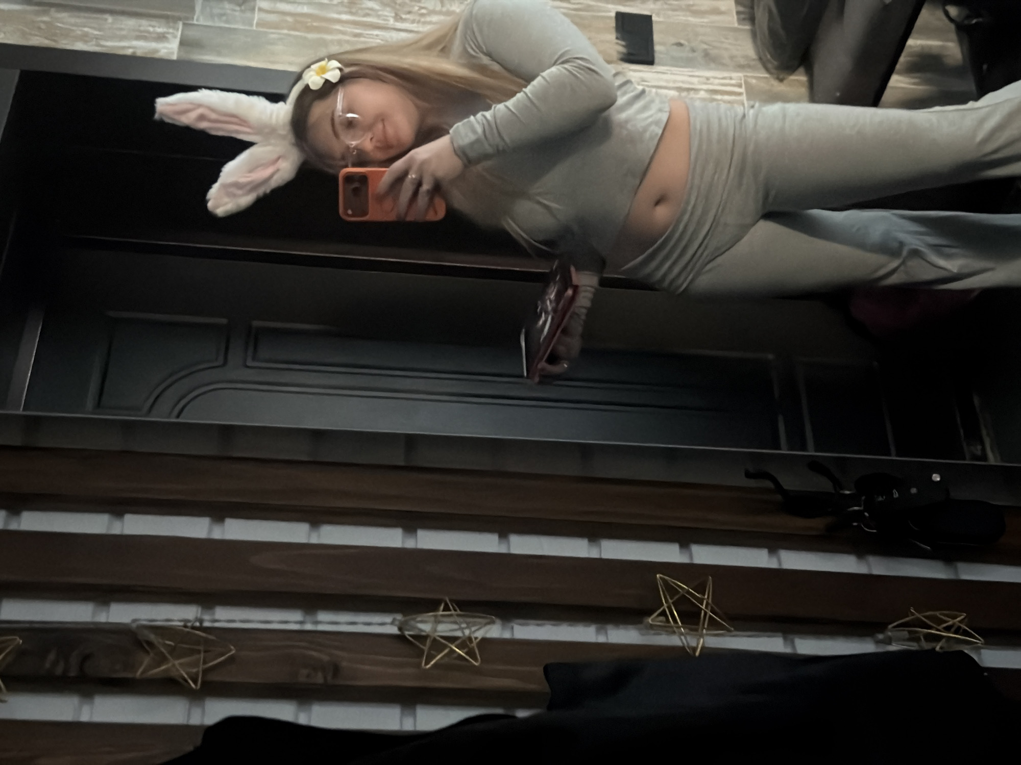 A person wearing a grey long-sleeve top and pants, with bunny ears and glasses, is taking a selfie in a mirror with an orange phone. The reflection includes a black door. The foreground contains a wooden plank wall decorated with gold star-shaped ornaments and a black object resembling a beetle. Location is unspecified.