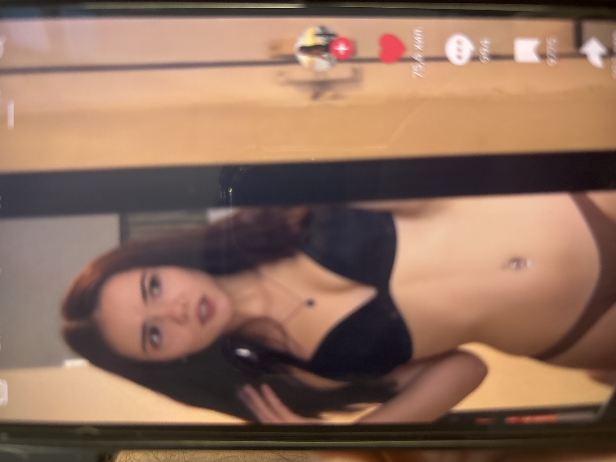 The image displays a photograph of a screen, likely a smartphone, showing a young woman in an indoor setting. The screen itself has visible borders and reflections.
The woman has long, dark brown hair parted in the middle. She is wearing a black push-up bra and dark brown underwear bottoms. A small, dark pendant on a thin chain is visible around her neck, and a shiny barbell-style navel piercing is present on her abdomen. Her face, while somewhat blurred, shows her looking towards the viewer's right, with her mouth slightly open, suggesting an expression of surprise or mild shock. Her right arm is slightly raised with her hand near her head/shoulder.
The background behind the woman consists of plain, light brown/tan walls, likely within a room or hallway, featuring vertical lines that may indicate door frames or wall panels. On the right side of the displayed content, social media interface elements are visible: a circular profile icon, a red heart icon followed by "75,6 млн." (75.6 million), a speech bubble icon with "994", a bookmark icon with "9775", and a share icon.
The woman is posing for the camera. The image does not contain any discernible information to specify the location as Blagoevgrad, Bulgaria, or any other specific geographic area; the environment is a generic indoor space.