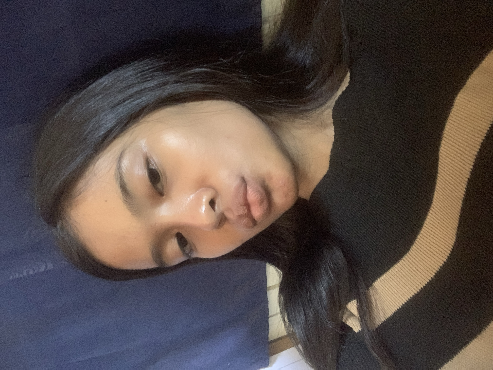 The media file displays a close-up portrait of a young woman, likely a selfie, taken indoors in what appears to be a personal space within Sungai Meriam, Indonesia. The woman is the primary subject, positioned centrally in the frame which is rotated, giving a tilted perspective. She has long, straight dark hair, dark eyebrows, and smooth skin with a noticeable sheen, particularly on her forehead and nose. Her lips are slightly pouted, suggesting a "duck face" or playful expression.
She is wearing a top that is predominantly black, featuring a prominent horizontal beige or light brown stripe across the right side of the garment. The background consists of a dark blue fabric, possibly a blanket, pillow, or upholstered furniture, on the left side of the frame, and a lighter, possibly off-white surface at the very bottom right, partially obscured by her hair and the blue fabric. The lighting appears soft and diffused, characteristic of indoor conditions.
The activity captured is a photographic pose. No specific event is evident beyond the act of taking a picture. Her expression is generally neutral to playful. There are no visible location cues within the image itself other than the provided context. No text is visible in the image or its crops.