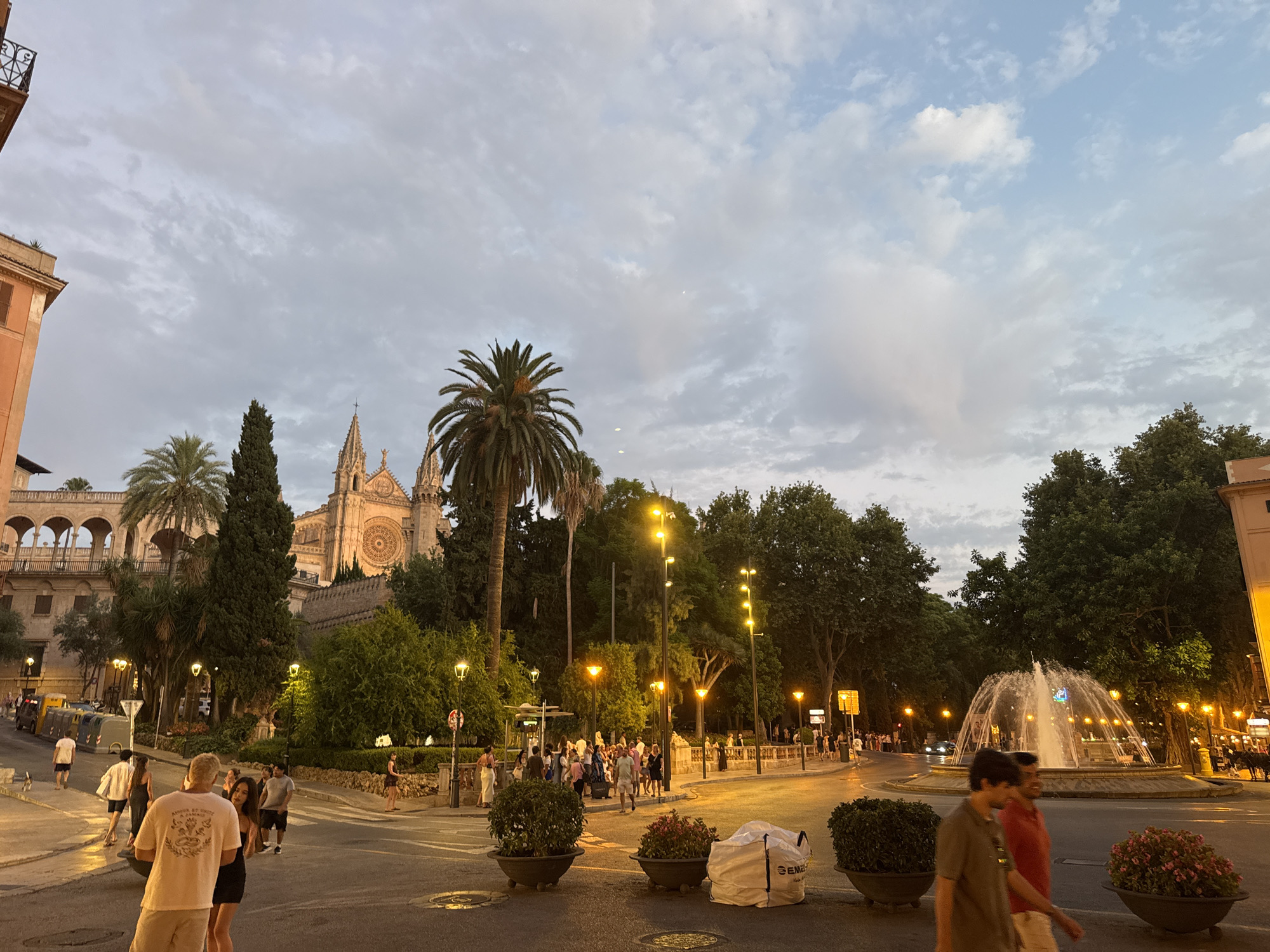 The image shows a street scene in Palma, Spain, featuring the Palma Cathedral in the background. The sky is overcast with a mixture of gray and light blue clouds. In the foreground, several people are walking on the street and sidewalk. To the right, a fountain is spraying water in a circular pattern. Streetlights are on, illuminating the scene with a warm glow. Trees and potted plants are visible along the sides of the street and around the fountain. The architecture suggests a European city with historical buildings, specifically the Gothic style of the cathedral.