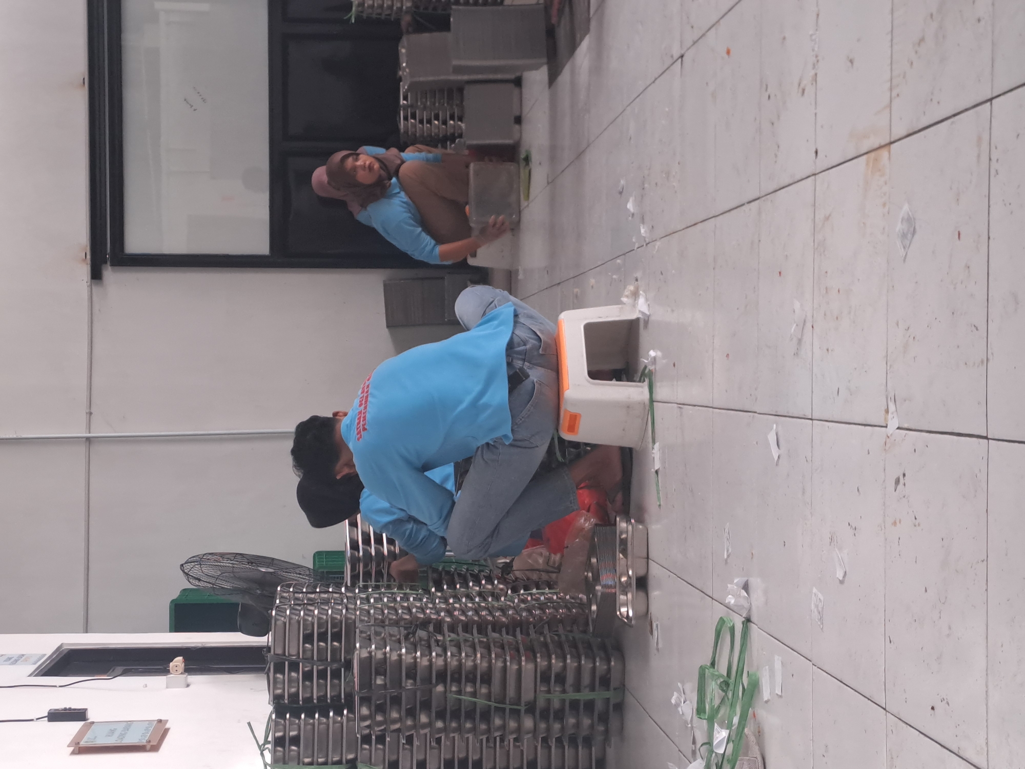 The image depicts an indoor scene with a tiled floor and white walls, possibly a factory or a large kitchen, located in Mojorejo, Indonesia. In the foreground, several stacks of metal trays are visible. Two individuals are partially visible, one in the middle ground and another in the background. The individual in the middle ground is wearing a blue long-sleeved shirt and jeans, sitting on a small white and orange stool, facing away from the camera. The individual in the background is wearing a light blue long-sleeved shirt and a headscarf, crouching down. The environment appears to be somewhat cluttered, with scattered white paper fragments and some green plastic packaging on the floor. A large industrial fan is visible in the mid-ground to the left. The overall impression is a utilitarian workspace.