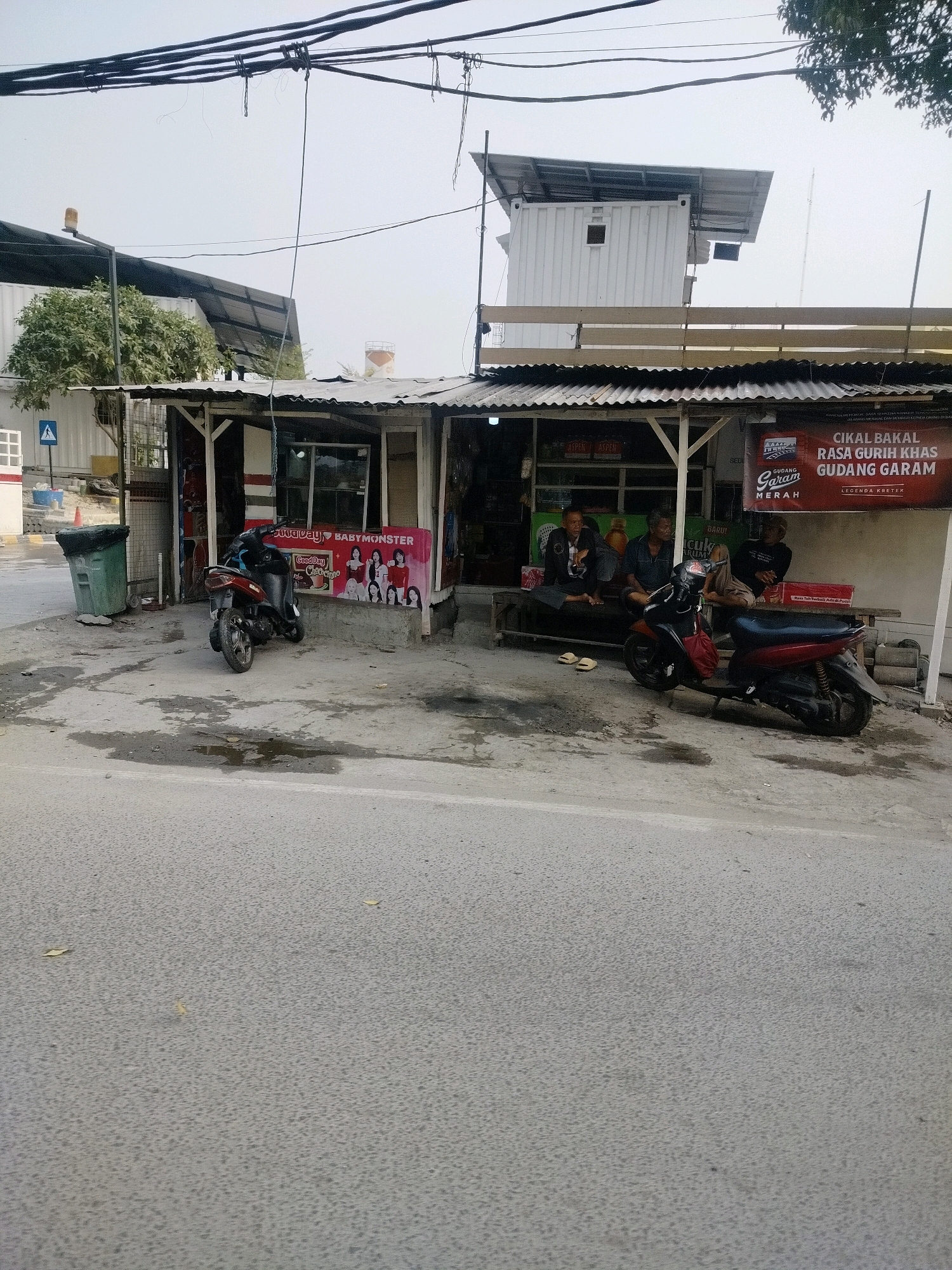 A street-level view captures a modest roadside shop or kiosk in East Jakarta, Indonesia, with a grey, overcast sky. The structure features a corrugated metal roof supported by wooden posts. In front of the shop, three men are seated on a wooden bench. Two of the men are engaged in conversation, while the third appears to be resting his head on his hand.
To the right of the shop, a red and black scooter is parked. A single red motorcycle is parked to the left of the shop, with its front wheel turned. A green trash bin with a black lid stands to the far left of the scene. In the background, a white shipping container structure with a metal roof is visible, possibly serving as an upper level or storage for the shop. Overhead, a dense network of electrical wires crisscrosses the sky. A sign with the words "GUDANG GARAM" and an image of what appears to be a cigarette pack is prominently displayed on the right side of the shop's facade. Another banner to the left features an image of a K-pop group, likely BabyMonster, and the words "Good Day". The ground in front of the shop is paved with concrete and shows signs of dampness with several puddles. A road with a textured asphalt surface occupies the foreground. A white sign with a blue pedestrian symbol is visible on a pole to the left of the shop.