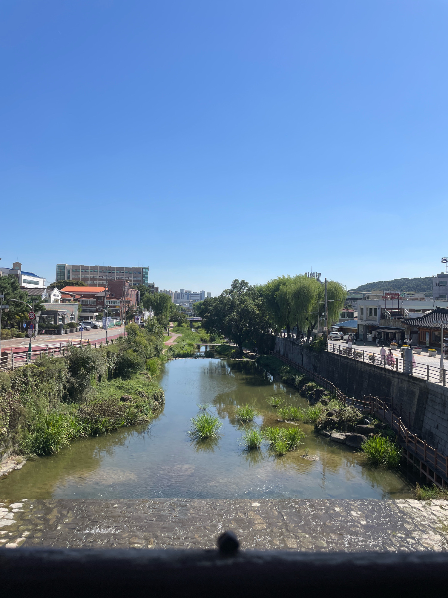 This is a landscape photograph taken on a clear day, looking down a canal or slow-moving river in Suwon-si, South Korea. The water is shallow and clear, with visible stones on the riverbed in the foreground and patches of green aquatic plants growing throughout. A small concrete dam or spillway is visible in the immediate foreground, creating a slight ripple in the water.
On the left bank of the river, there is a well-maintained walking path bordered by greenery and trees. Beyond the path, there are buildings of varying heights and architectural styles, including a multi-story brick building with an orange roof and a taller, modern building in the distance. A road with traffic signs and parked cars runs parallel to the path.
The right bank of the river features a stone wall with a wooden railing and walkway. Several people are visible walking along this path, some dressed in traditional Korean clothing, suggesting a cultural or historical area. Further along, there are more buildings, some with traditional tiled roofs, and lush green trees, including weeping willows. In the distance, rolling hills and more urban development can be seen under a bright blue sky. The overall impression is a peaceful urban scene blending nature with the cityscape.