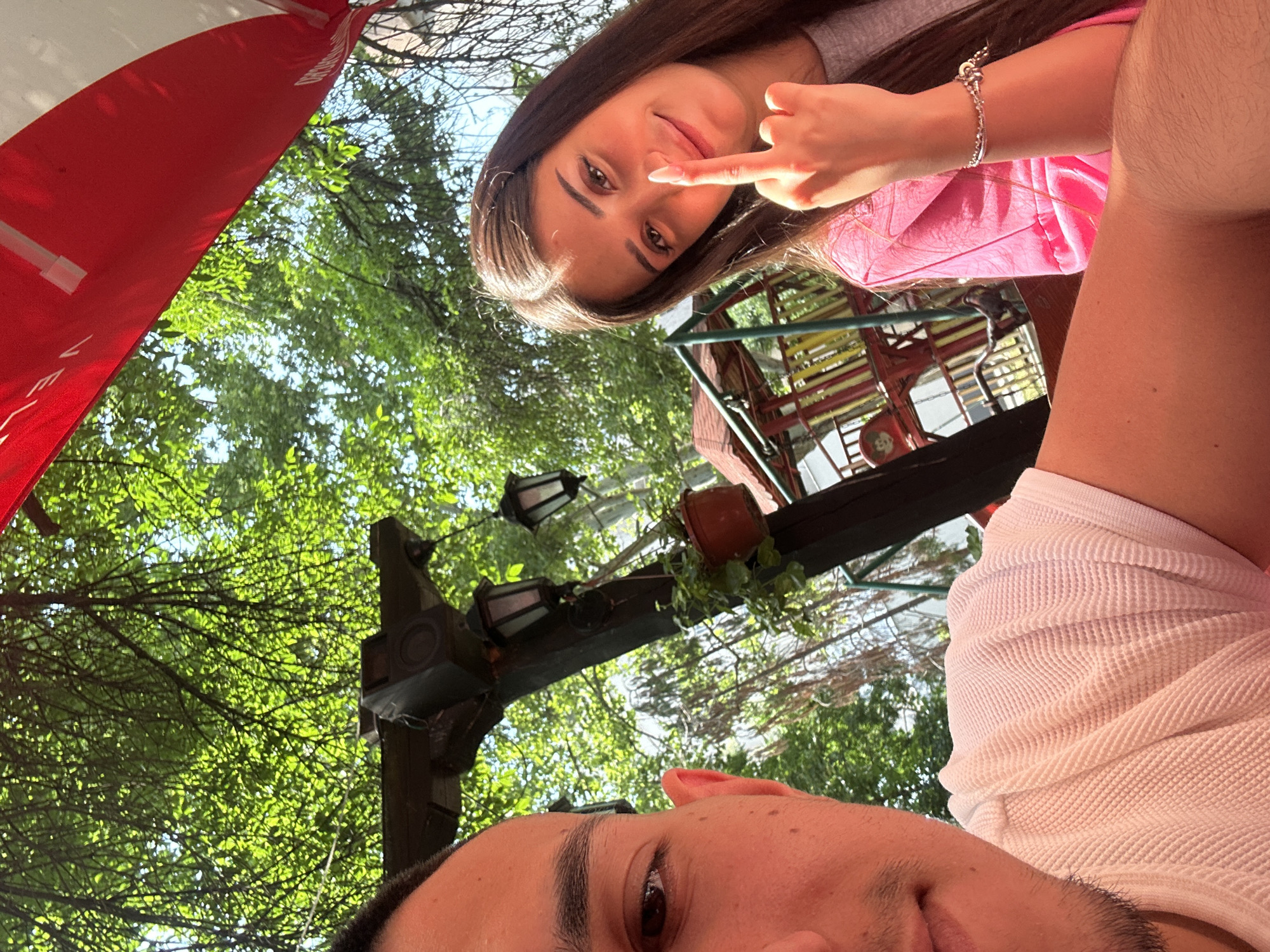 The image is an outdoor selfie taken from a low angle, featuring two individuals with lush green foliage and architectural elements in the background. The photograph is oriented so the subjects appear sideways.
In the upper-middle section, a young woman with long, dark brown hair looks directly at the camera with a subtle smile. She wears a grey t-shirt over a bright pink top or dress, and a silver bracelet is visible on her left wrist. Her right index finger is playfully touching her nose.
The lower part of the frame shows the top of a man's head, including his right eye, eyebrow, and part of his cheek and ear. He is wearing a white, textured shirt, possibly with a waffle knit pattern.
The background features abundant bright green tree leaves, suggesting a sunny daytime setting. Above the man's head, dark wooden beams support decorative hanging lanterns and a small potted plant, indicating an outdoor patio or garden area, consistent with a cafe or restaurant in Plovdiv, Bulgaria. On the far left, a red and white striped awning or umbrella is partially visible, bearing white text that reads "VELO."
The bright natural lighting confirms it is daytime. The playful gesture and relaxed expressions convey a cheerful and informal atmosphere.
