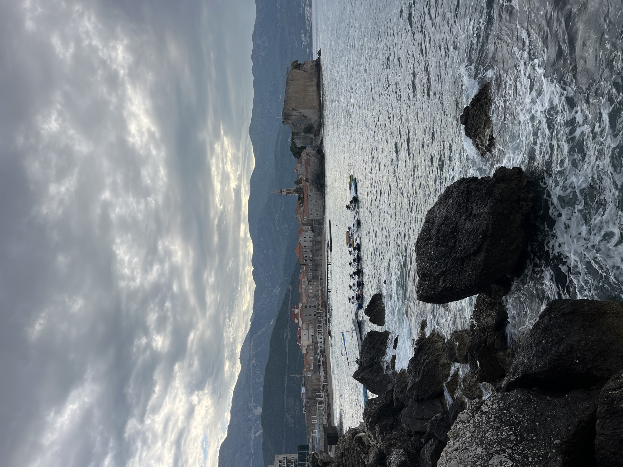 The image captures a dramatic coastal landscape, presented in a portrait orientation. The left side is dominated by a cloudy sky, filled with dense, grey and white clouds, where patches of brighter light hint at sunlight breaking through.
Towards the center and extending to the lower right, a rugged coastline unfolds. A historic town, characterized by old stone buildings and a prominent bell tower, is nestled at the base of a towering mountain range. This town is built on or around a rocky promontory that juts into the sea, featuring an ancient-looking fort or citadel on its higher reaches. Several small boats and jet skis are moored in the choppy waters near the town's edge.
In the far right and lower foreground, dark, craggy rocks are visible, against which white-capped waves crash, creating foamy patterns on the dark grey surface of the sea. The sea occupies a significant portion of the right side of the frame, appearing turbulent. The mountains rise steeply behind the town, their slopes covered in sparse vegetation, extending upwards towards the cloudy sky on the far left.
The overall lighting is subdued, suggesting either an early morning, late afternoon, or a heavily overcast day. The scene evokes a sense of rugged natural beauty combined with historical architecture, consistent with a location like Budva, Montenegro. There is no discernible text visible in the image.