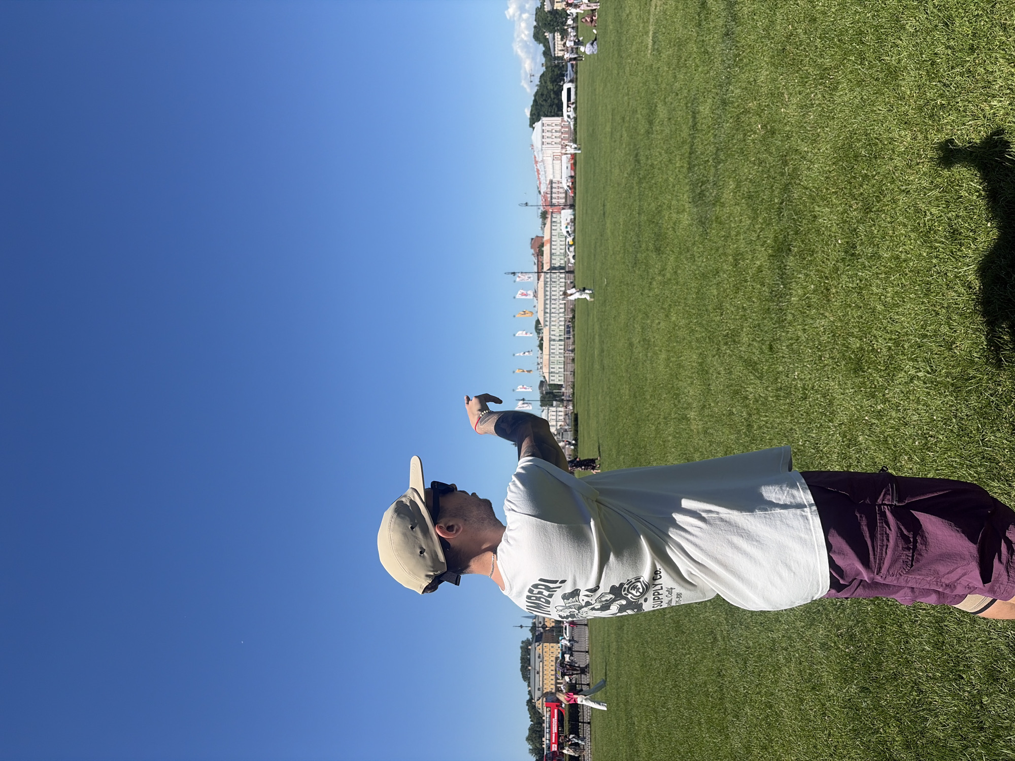 A man wearing a light-colored t-shirt, burgundy cargo shorts, a tan baseball cap, and sunglasses is standing in a large grassy field. He is facing away from the camera and pointing his left hand towards a building in the distance. The man has tattoos on his left arm. In the background, there are several buildings with flags, some people walking and sitting on the grass, and a red double-decker bus. The sky is clear and blue. This scene is likely located in Palace Square in Saint Petersburg, Russia, a prominent historical and architectural complex in the city.
