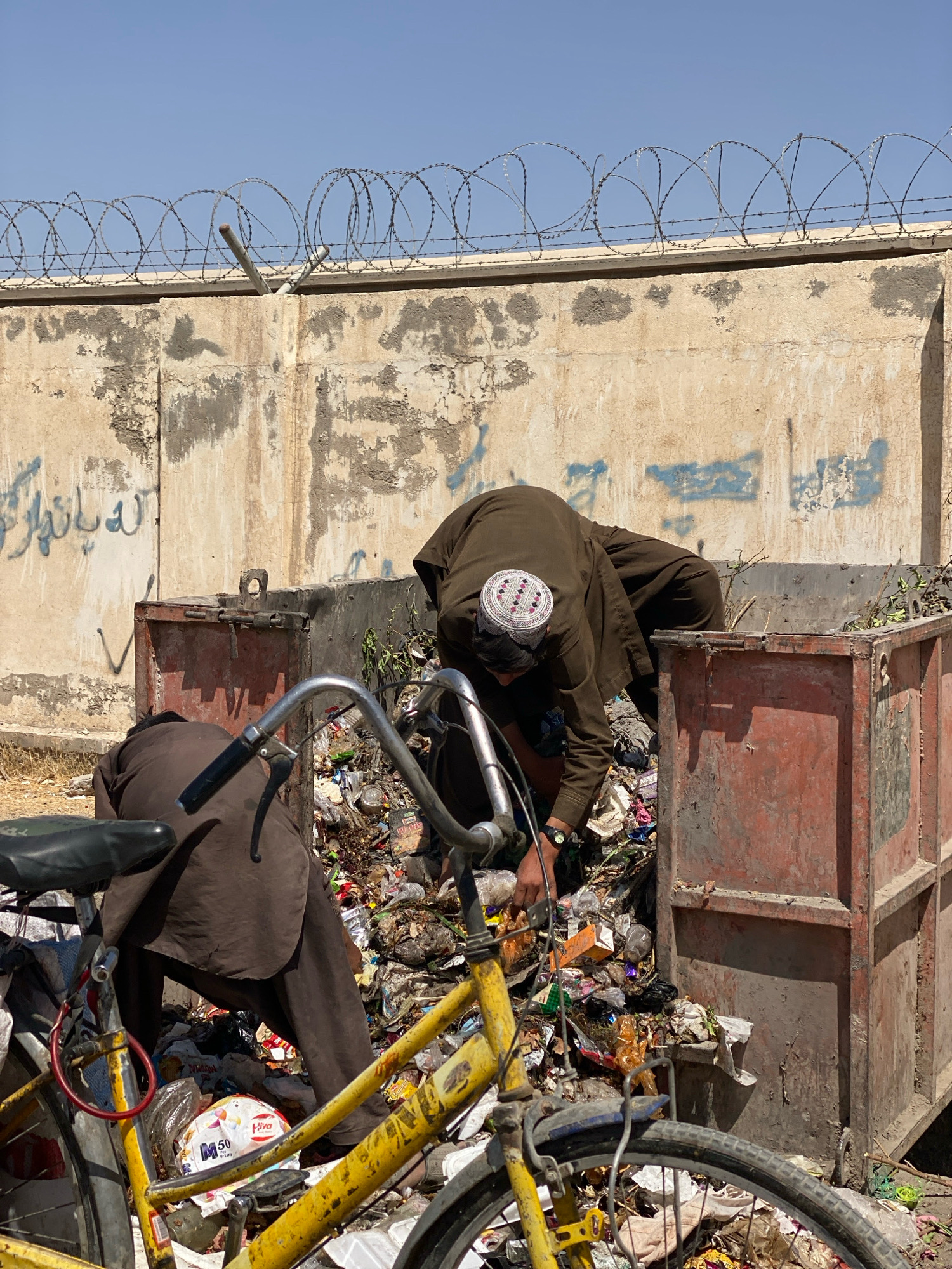 In Kandahar, Afghanistan, two individuals dressed in traditional brown clothing are sorting through a large, rusty metal dumpster overflowing with garbage. One person, wearing a white and red patterned cap, is bent over inside the dumpster, actively sifting through the refuse. The other person, partially visible, is crouched down beside the dumpster, also appearing to be engaged in the same activity. A yellow bicycle is positioned in the foreground, partially obscuring the dumpster and the individuals. The backdrop consists of a weathered concrete wall topped with barbed wire, and the ground is covered with dry grass and debris. The sky is clear and blue.