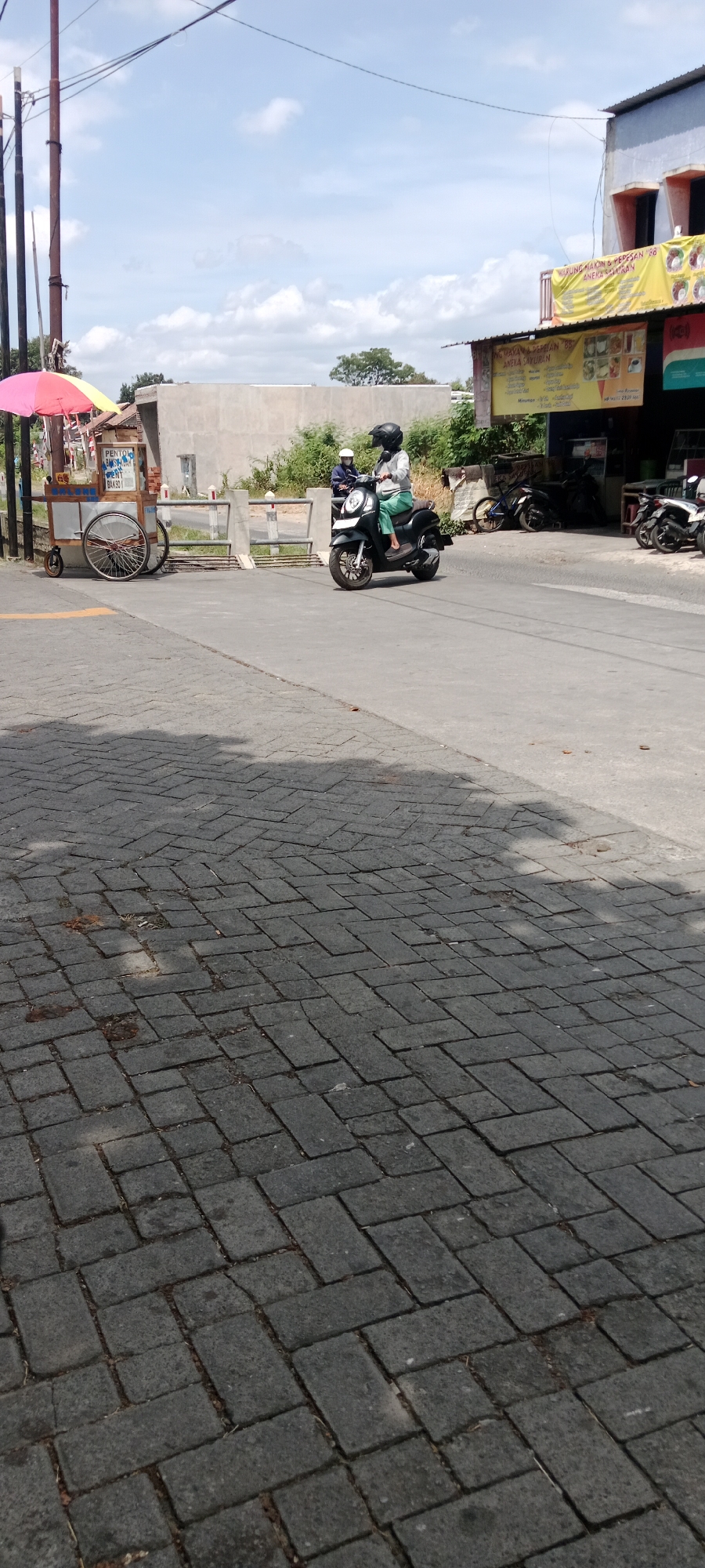 In Maguwoharjo, Indonesia, a dark-colored scooter is being ridden by a person wearing a helmet and a light-colored jacket over green pants. The rider is moving on a paved road, which is bordered by a sidewalk made of dark rectangular bricks. To the left of the road, there is a food cart with a colorful umbrella, a wheeled cart, and signage that appears to advertise food items. Behind the food cart and across the road, a light-colored building with a corrugated metal roof stands. In the background, there is a concrete wall and some vegetation. The sky is partly cloudy. Several other motorcycles are parked in front of the building.